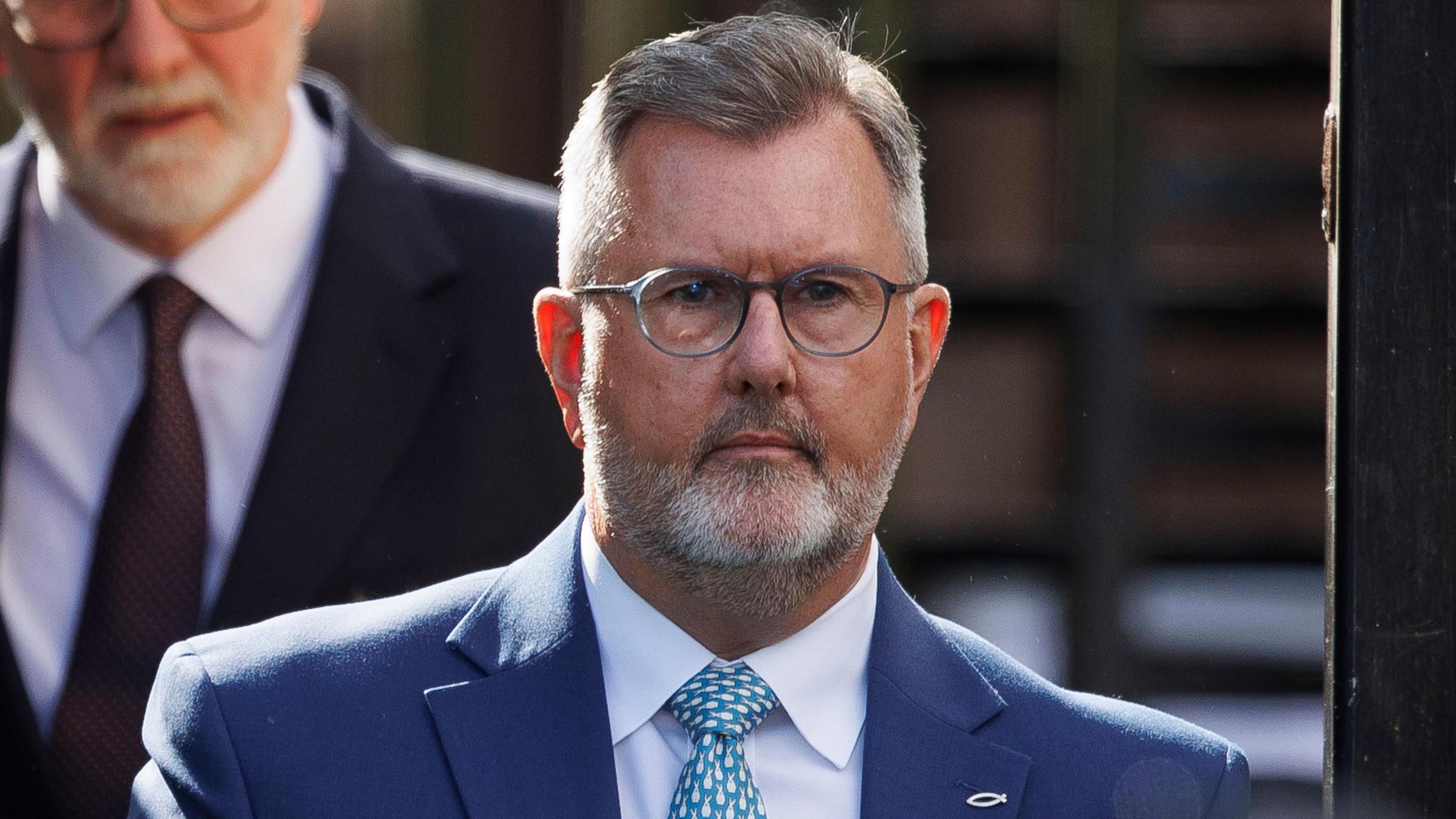 Sir Jeffrey Donaldson - a man with short, grey hair and a beard wears silver, circle glasses. He is wearing a blue suit jacket, a white collared shirt and a light blue patterned tie.