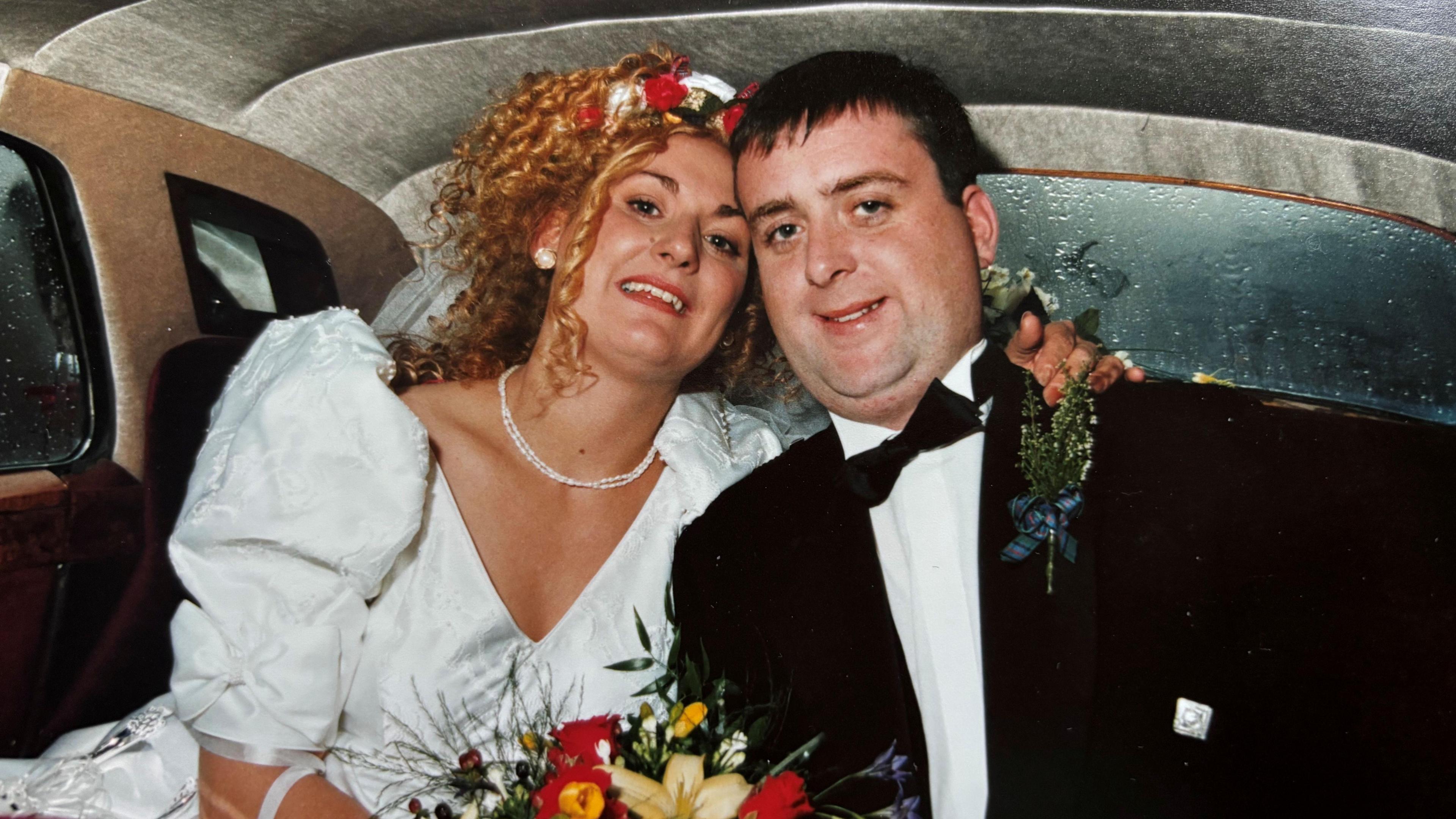A newly wed couple just after their marriage, sitting smiling in the back of a car. The bride is wearing a white wedding dress and has strawberry blonde hair - the groom is wearing a suit and has short dark hair.