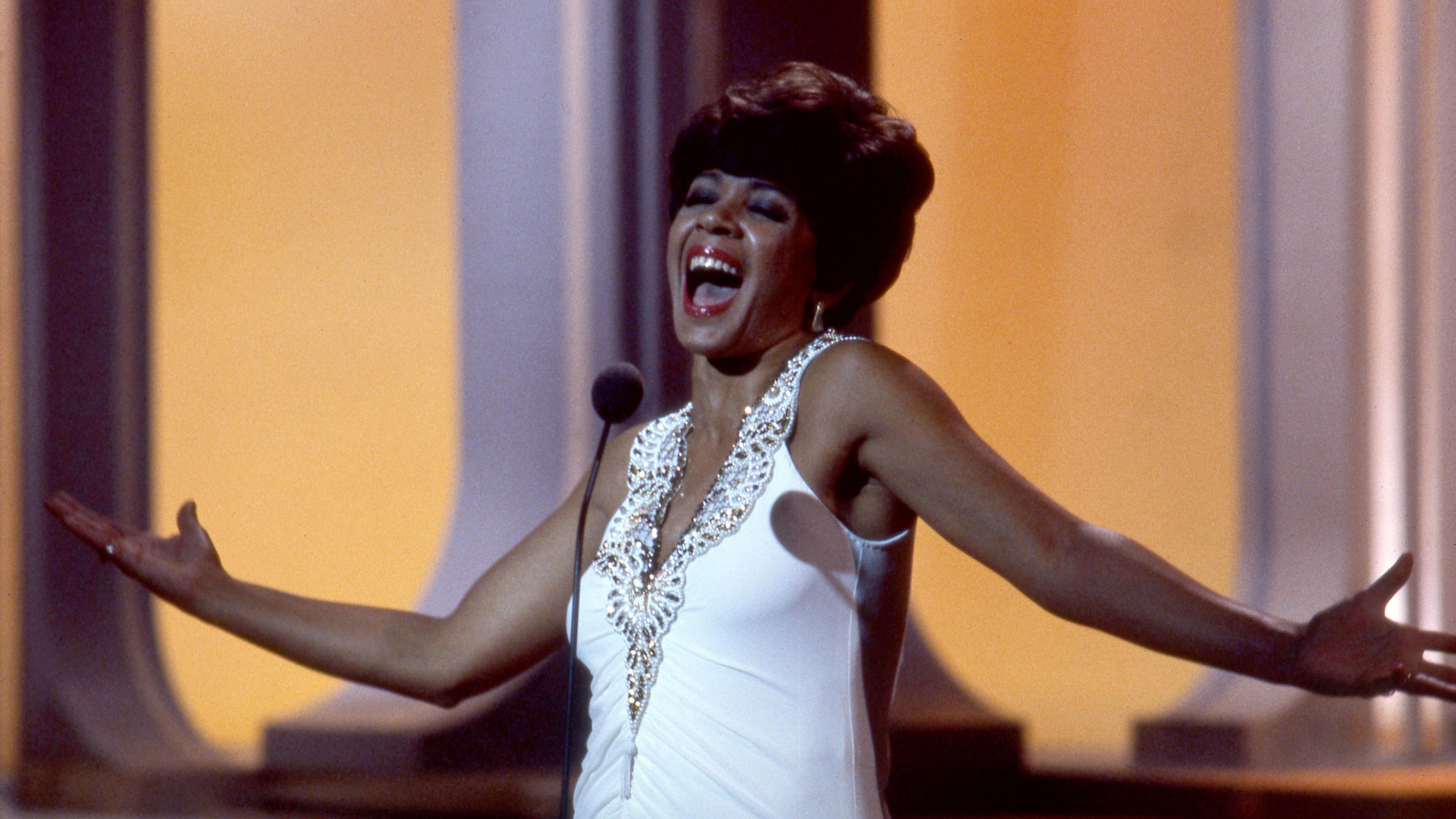 Shirley Bassey wearing a white dress while singing