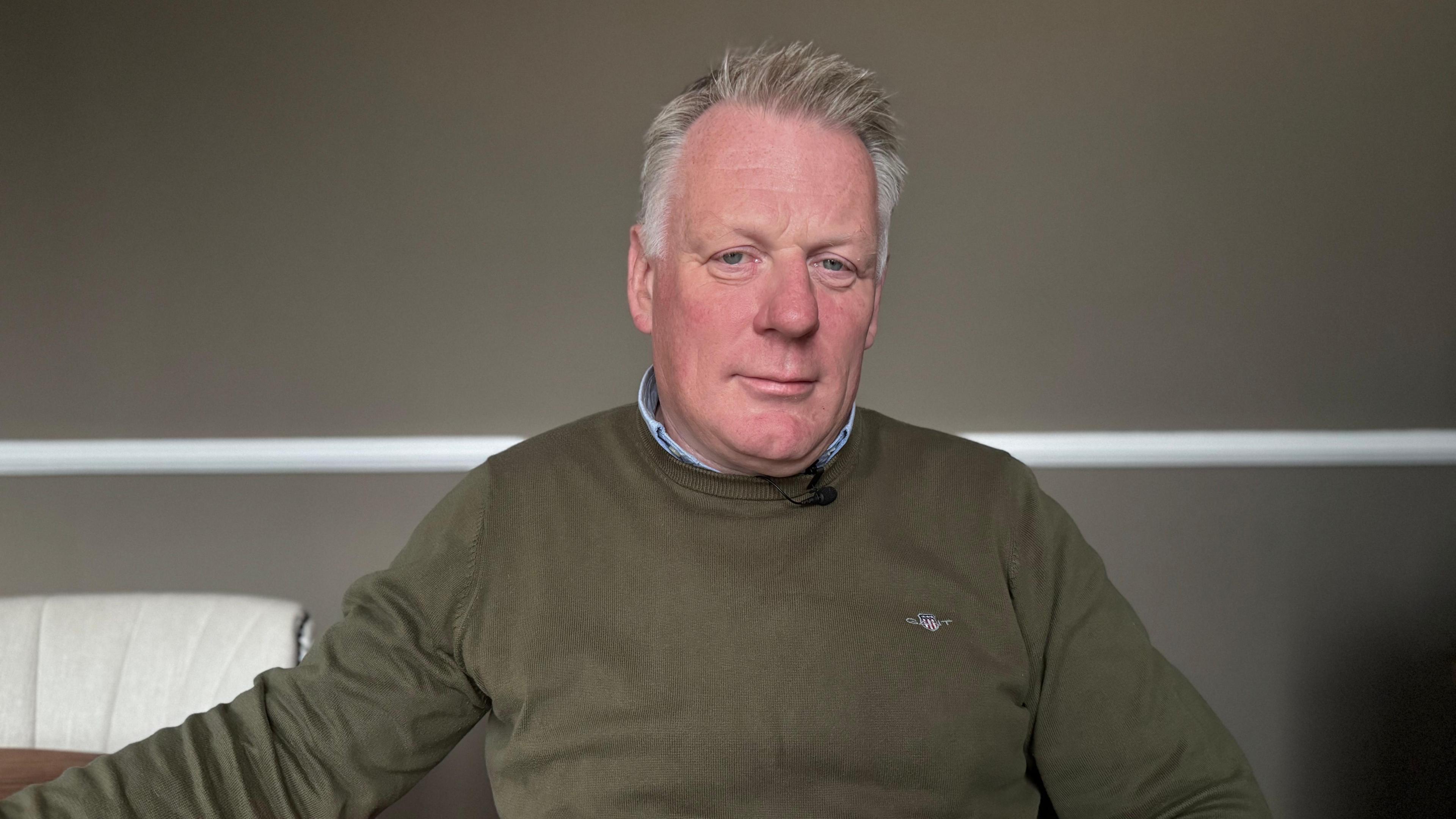 Man with grey hair looking into camera, wearing green/brown round neck jump