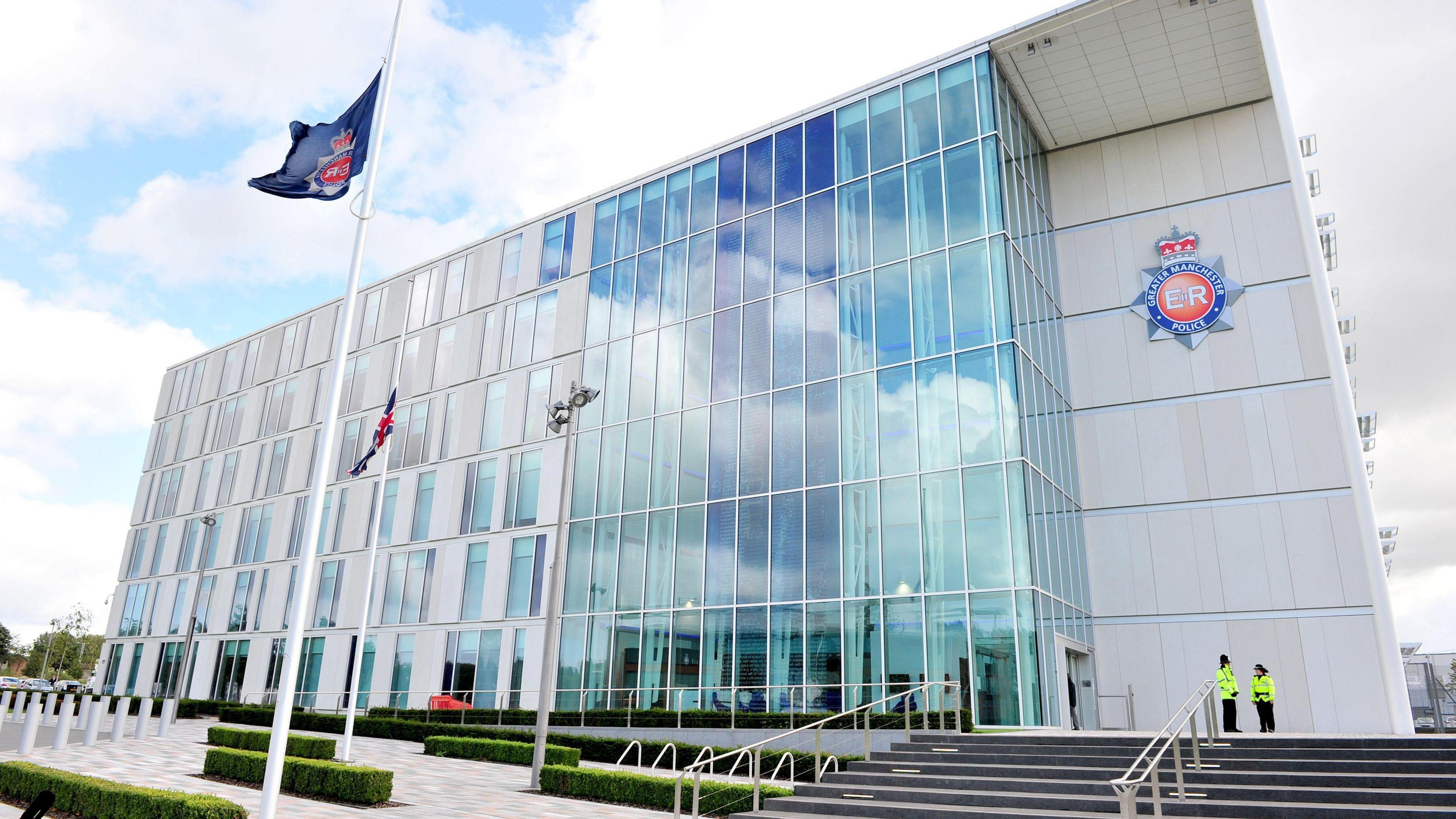 Greater Manchester's glass-fronted force headquarters, against a blue sky with two officers and a flag in the foreground