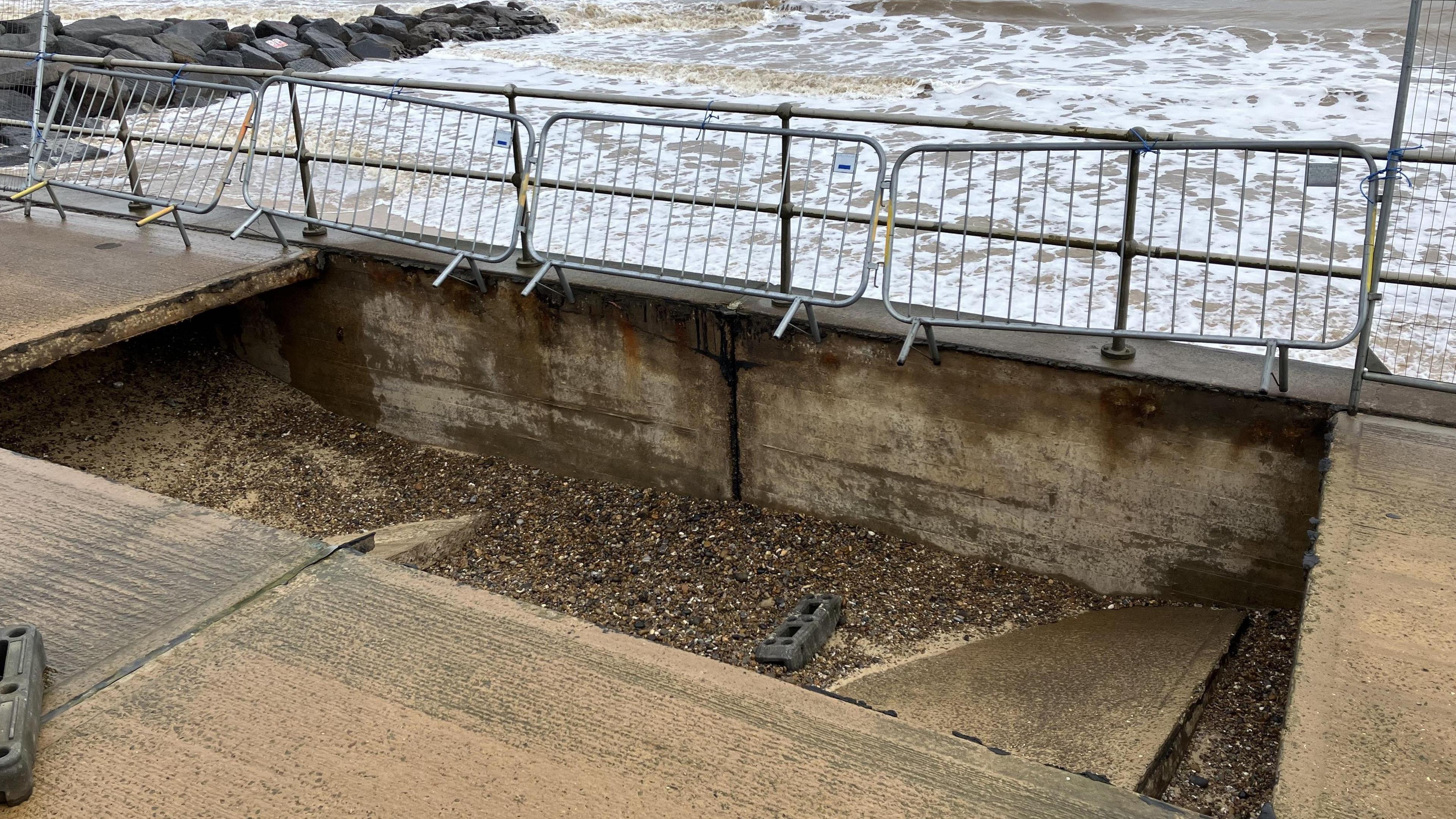 Metal fencing is in background and a large, rectangular-shaped hole in a concrete promenade has shingle inside and the remnants of the collapsed walkway.