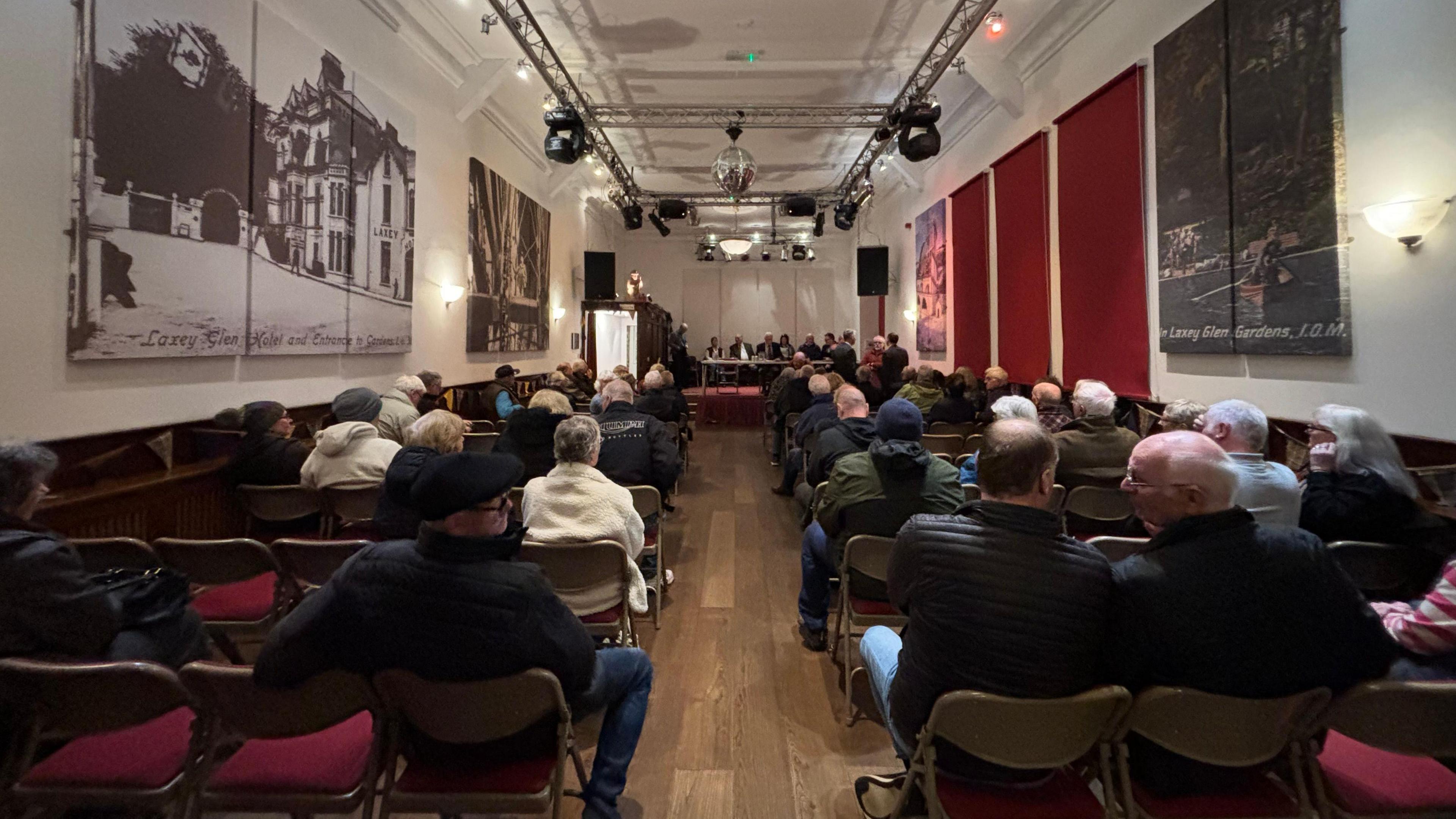 People in a hall sitting on chairs with red seats. It is a long hall with a wooden floor and white walls.