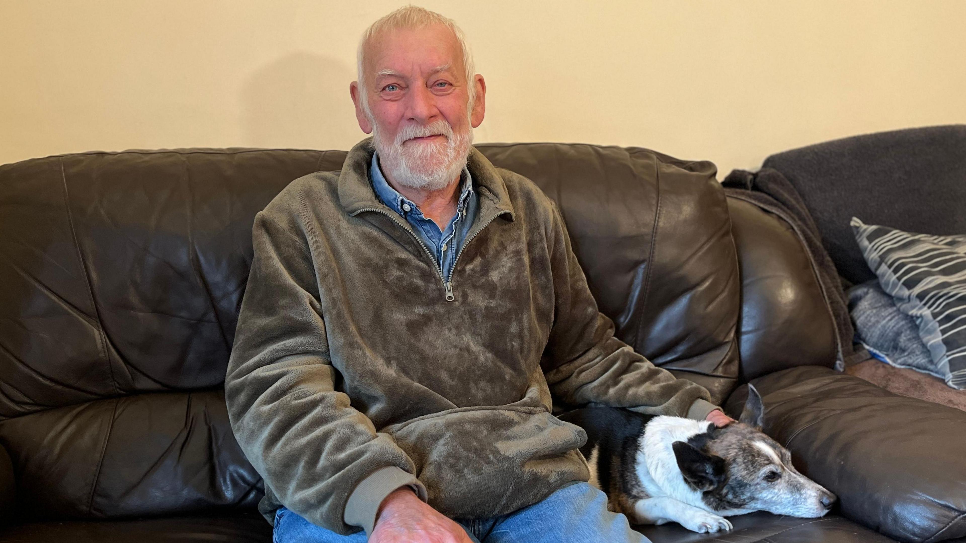 A man with short grey hair and beard is sitting at home on a brown leather sofa next to his small black and white dog. He is wearing a light brown hoodie and has his hand on his dog.