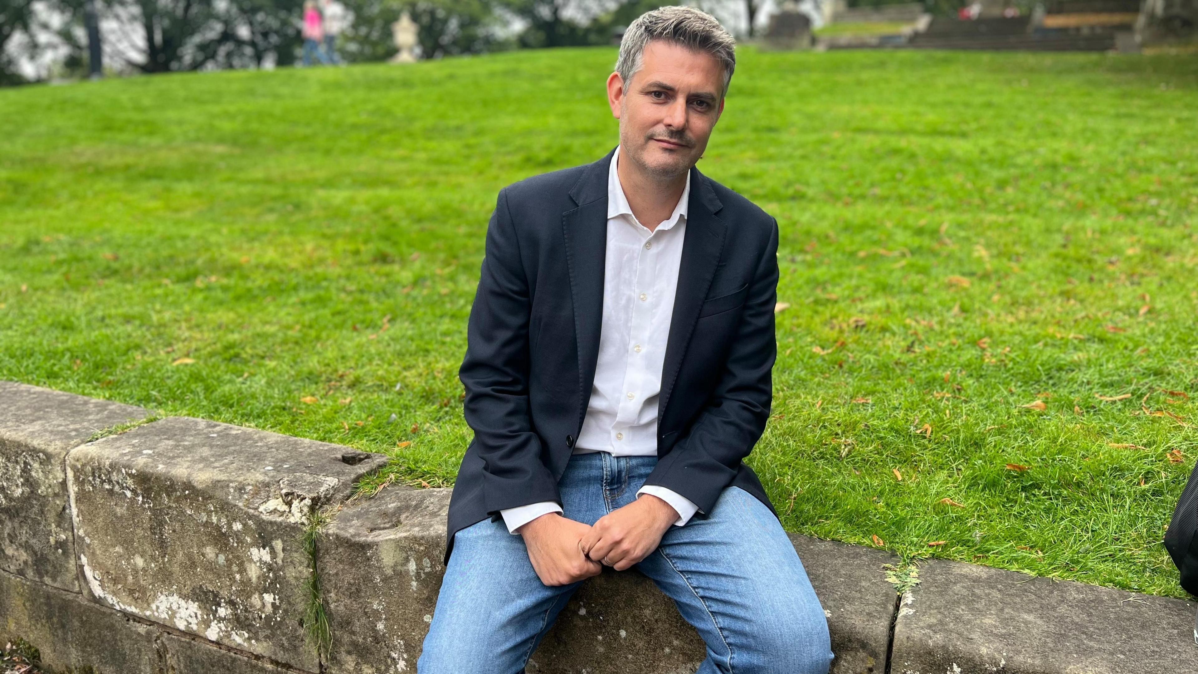 Man with short grey hair in white shirt navy blazer blue jeans sat on stone wall infront of grass verge 
