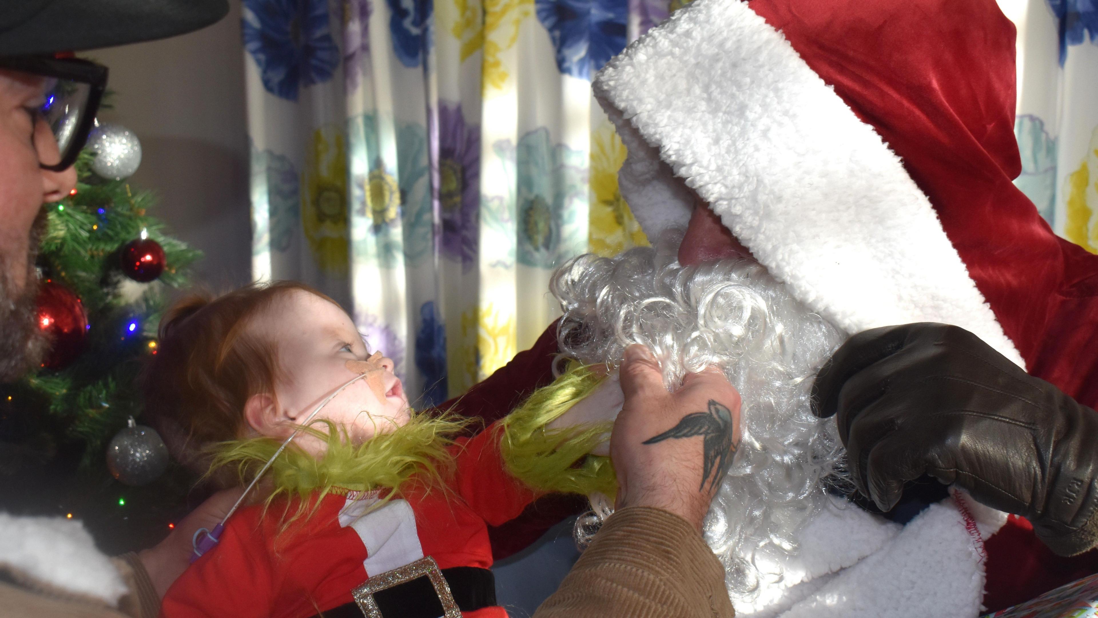 A man dressed as Father Christmas stares down at a toddler at Children's Hospice South West in Wraxall. The child, who is being supported by their father, is also wearing a Christmas-style red and white and black outfit.