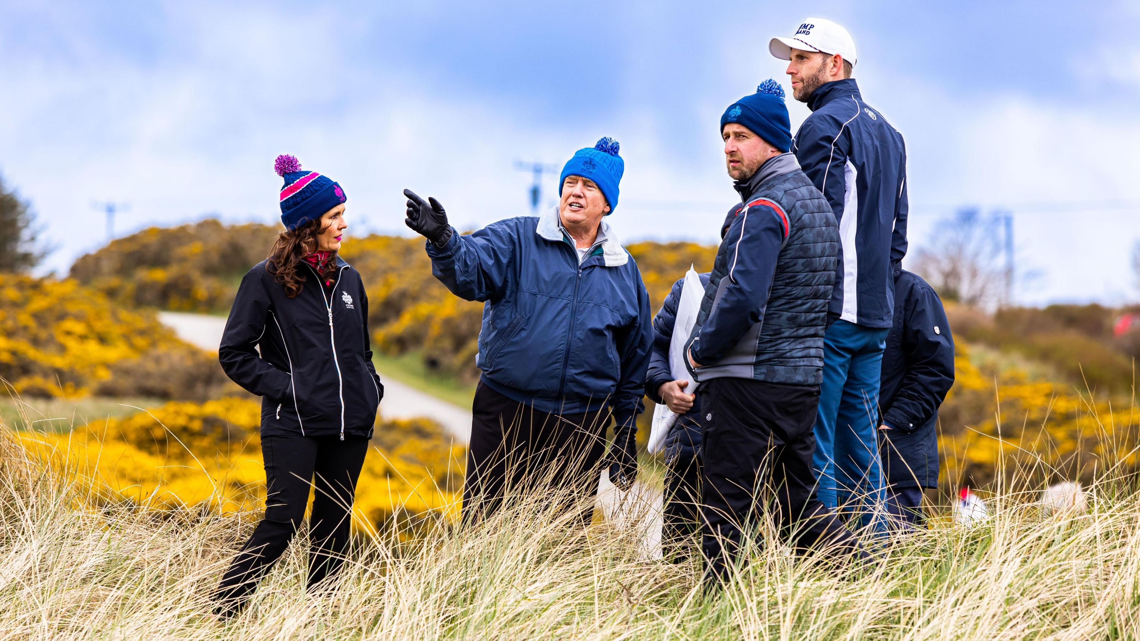 Donald Trump at the site of his new course near Menie in Aberdeenshire. He is standing in long grass wearing a blue jacket and a blue bobble hat. A group of people also wearing dark jackets and hats surround him.