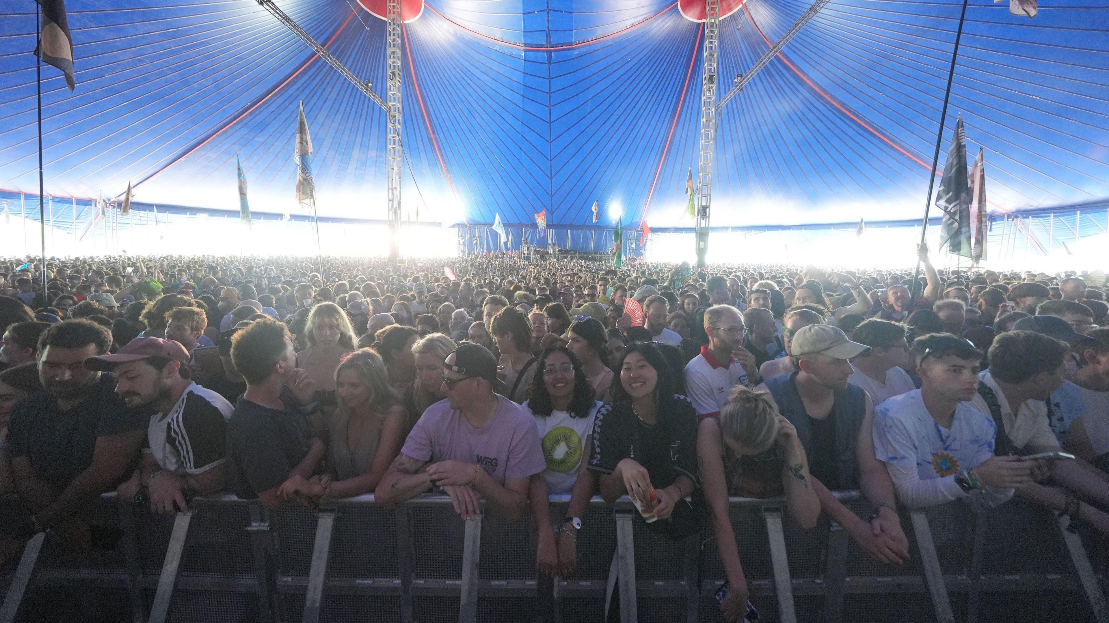 A crowd gathering by a stage seen from the side of the stage. They are in a blue tent.