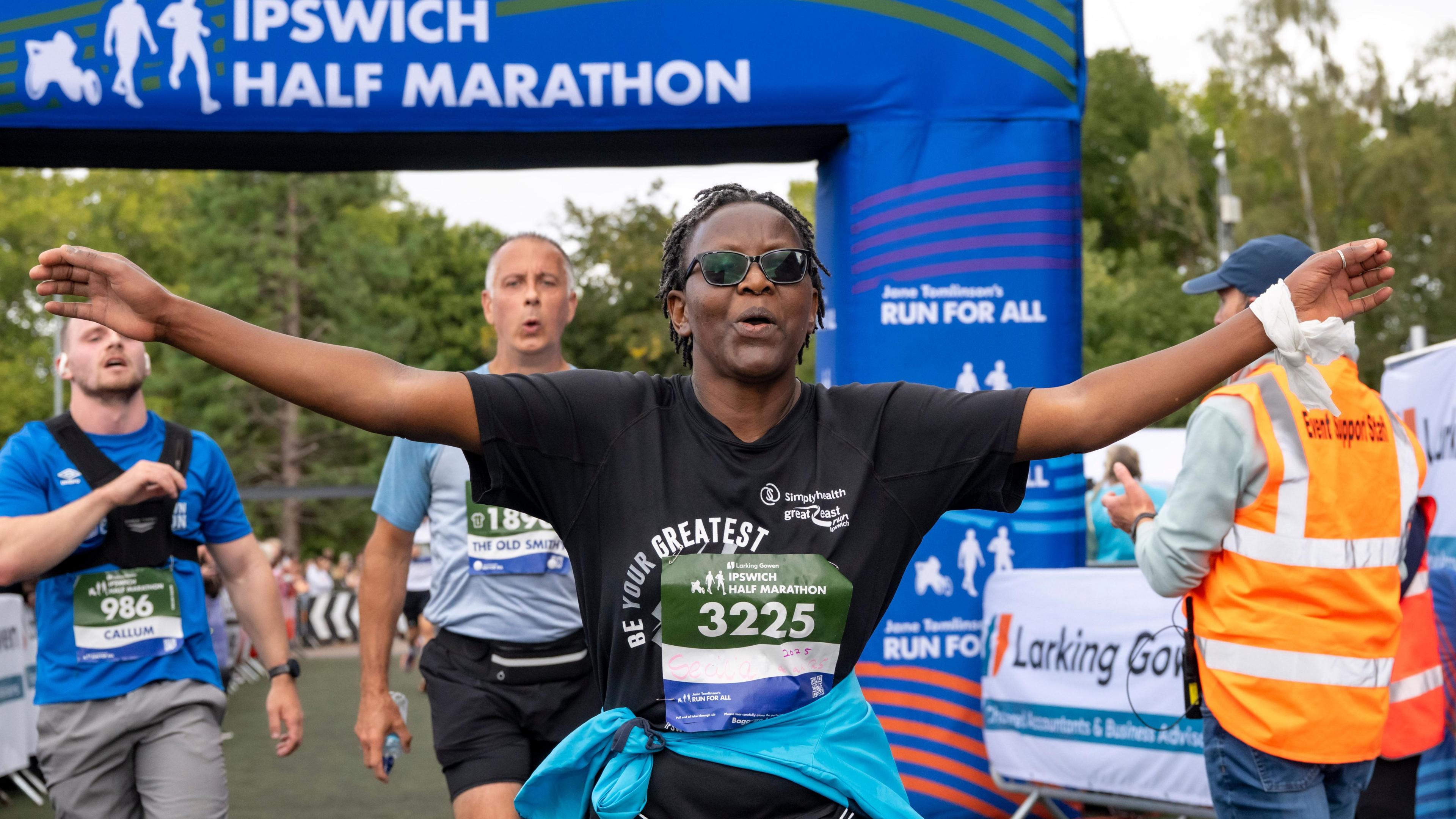 A man dressed in black has his arms outstretched as he passes under the finishing arch at the marathon, other exhausted runners behind him.