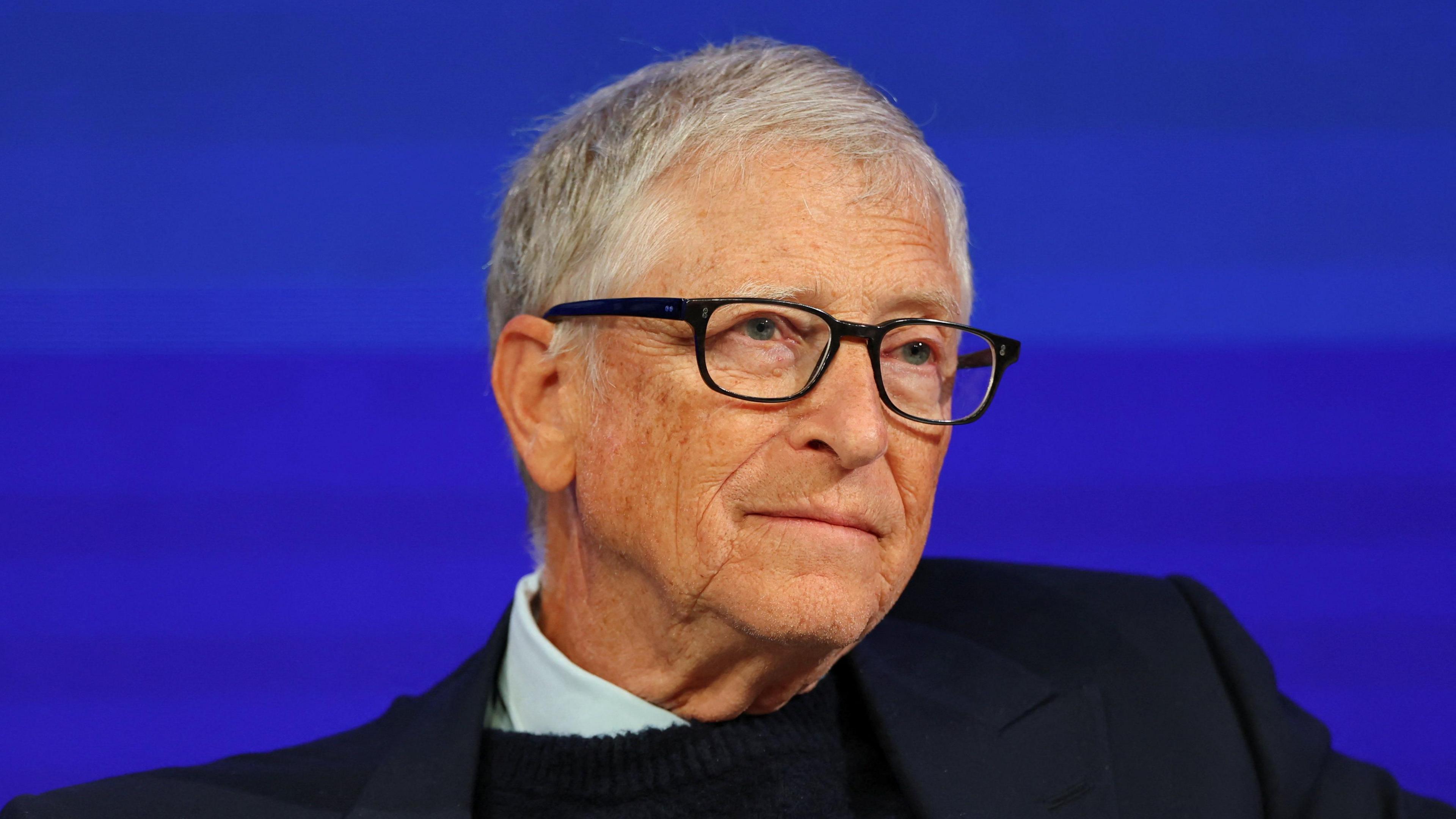 Bill Gates looks away from the camera with a blank expression. He is sat in front of a blue backdrop on-stage at the World Economic Forum in Davos.
