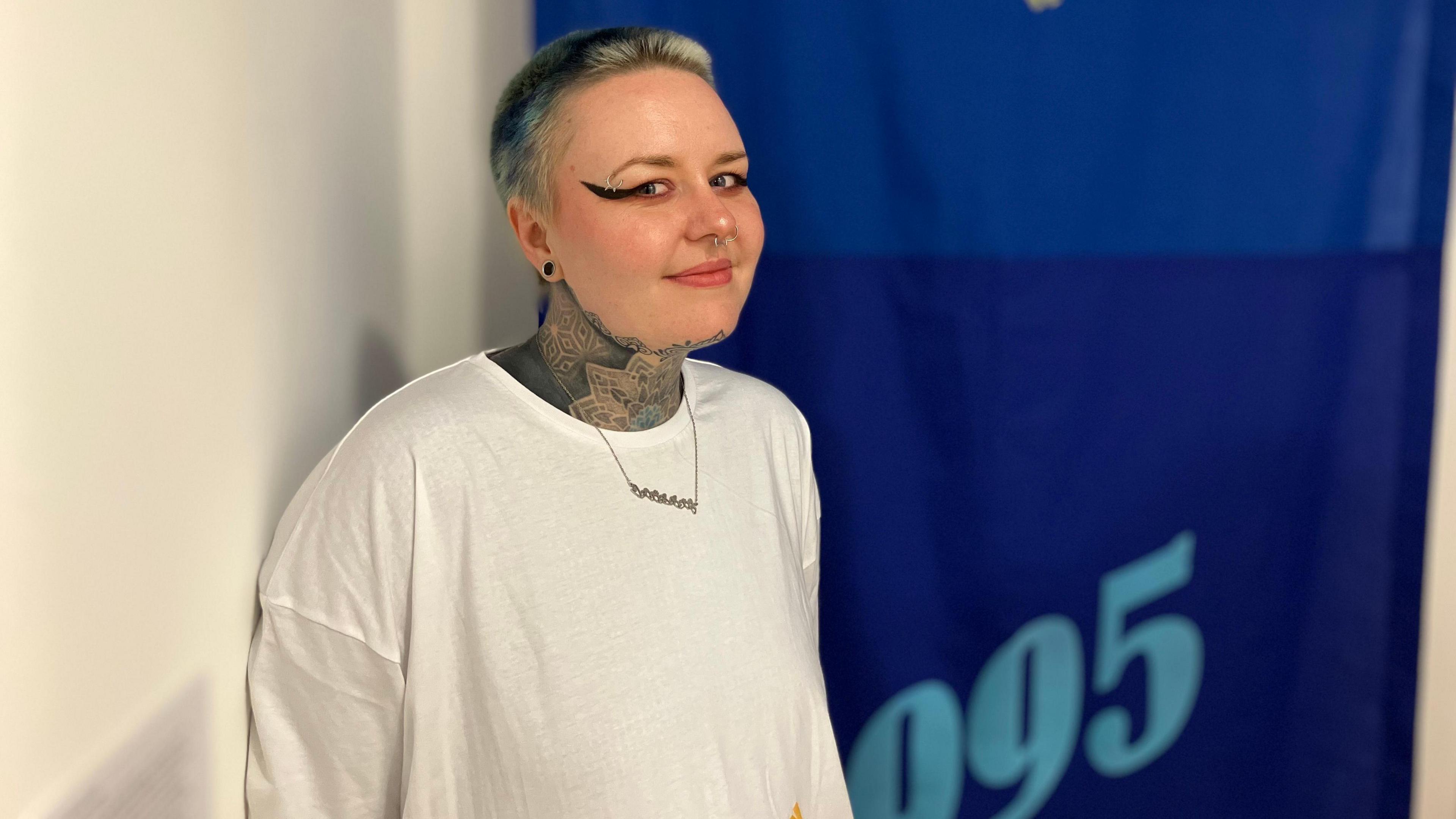 Charley wearing a white shirt smiling for the camera standing in front of a blue 1996 banner. She has a heavily tattoed neck