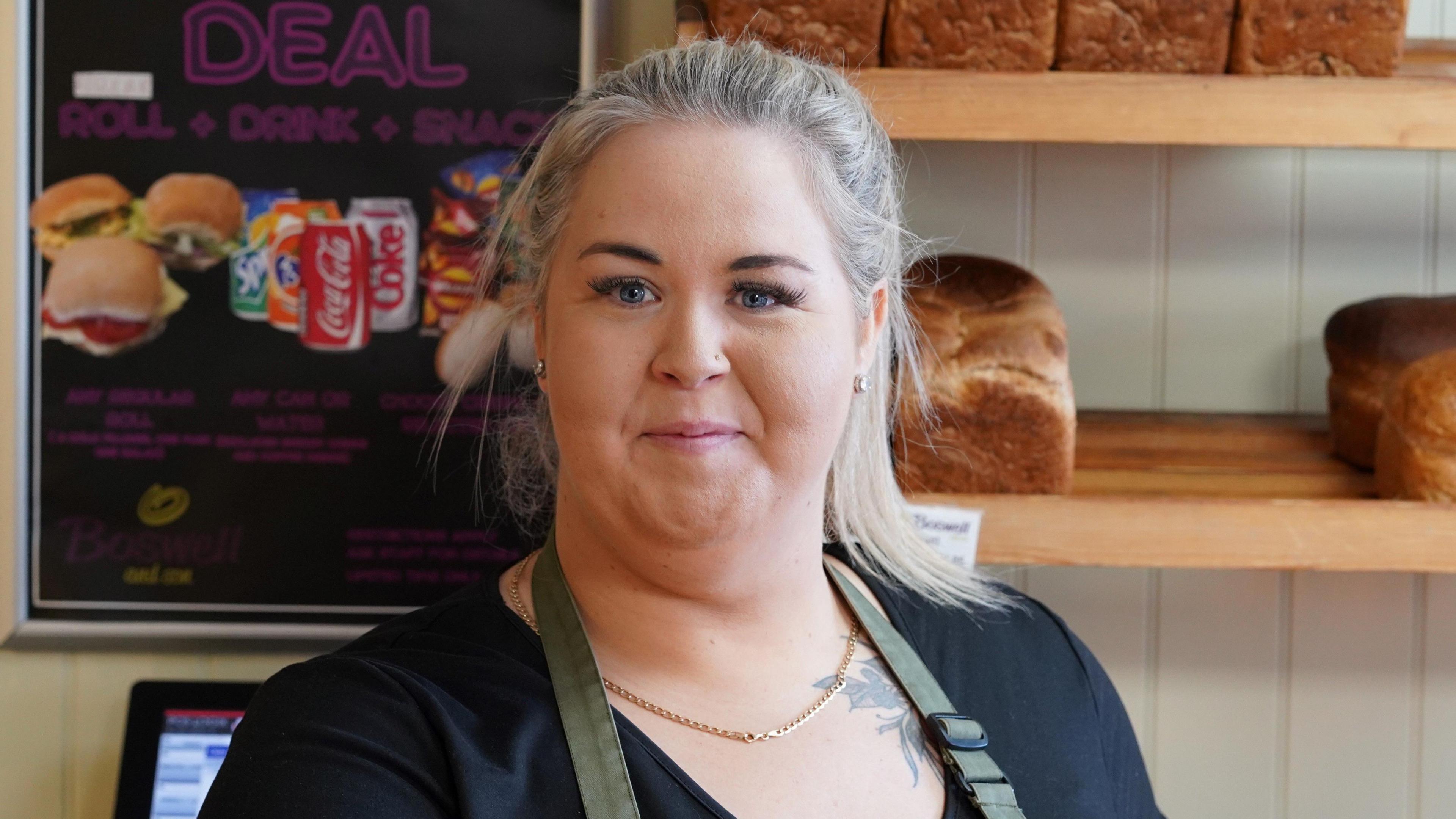 Louise is wearing an apron and standing behind the bakery counter, behind are are shelves with bread loaves on them.