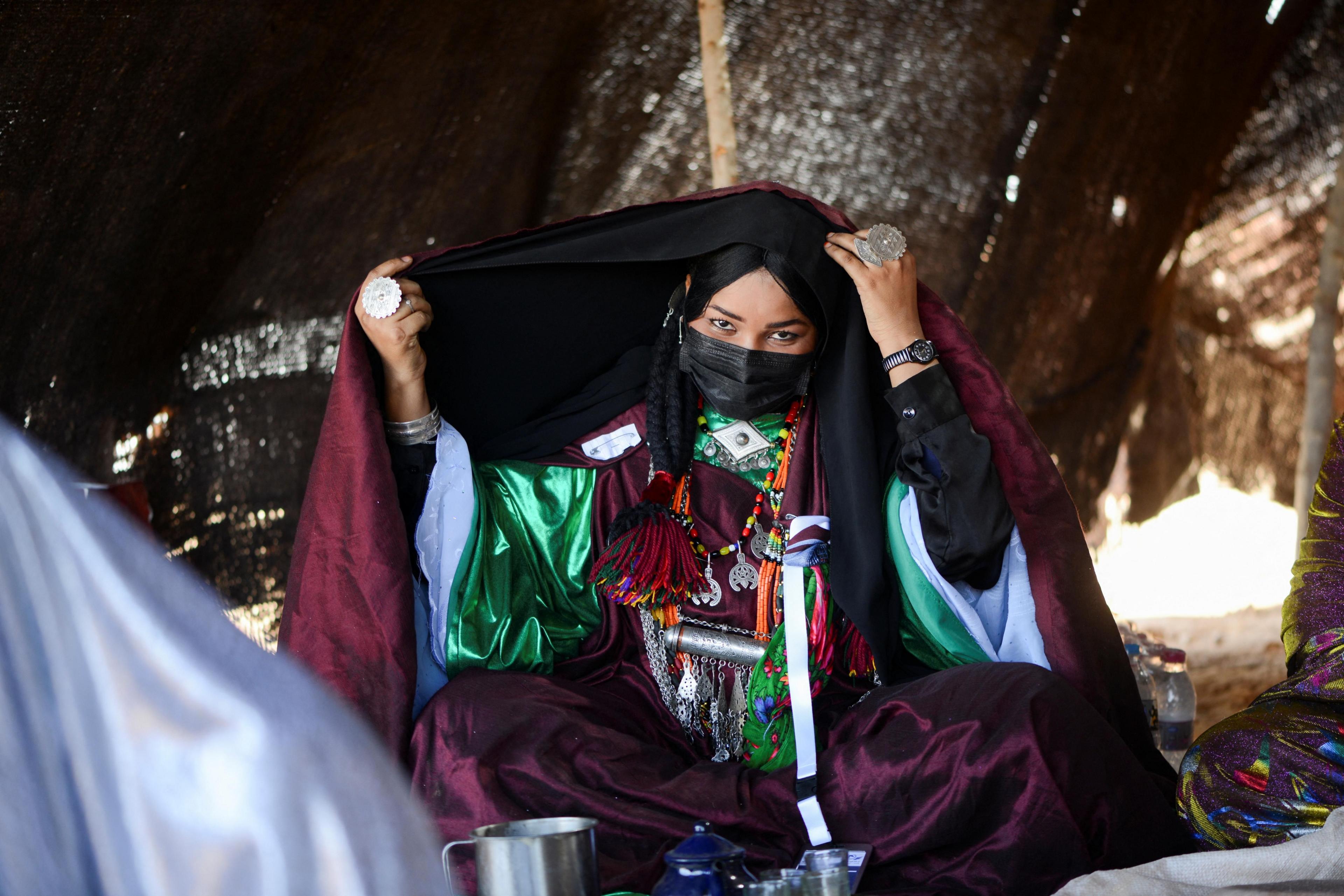 A woman sits cross-legged in a desert tent. wears plum coloured robes and bold silver jewellery.