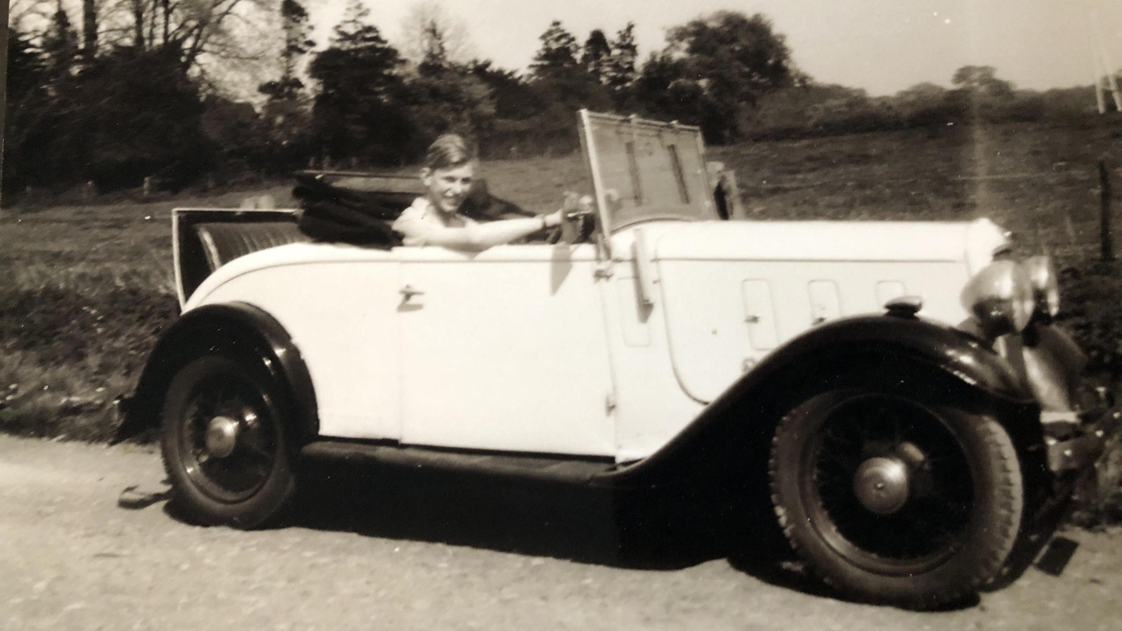 An old black and white photograph of a teenager smiling while sitting in a white old car with the roof down. Behind the car is a field with trees on the horizon.