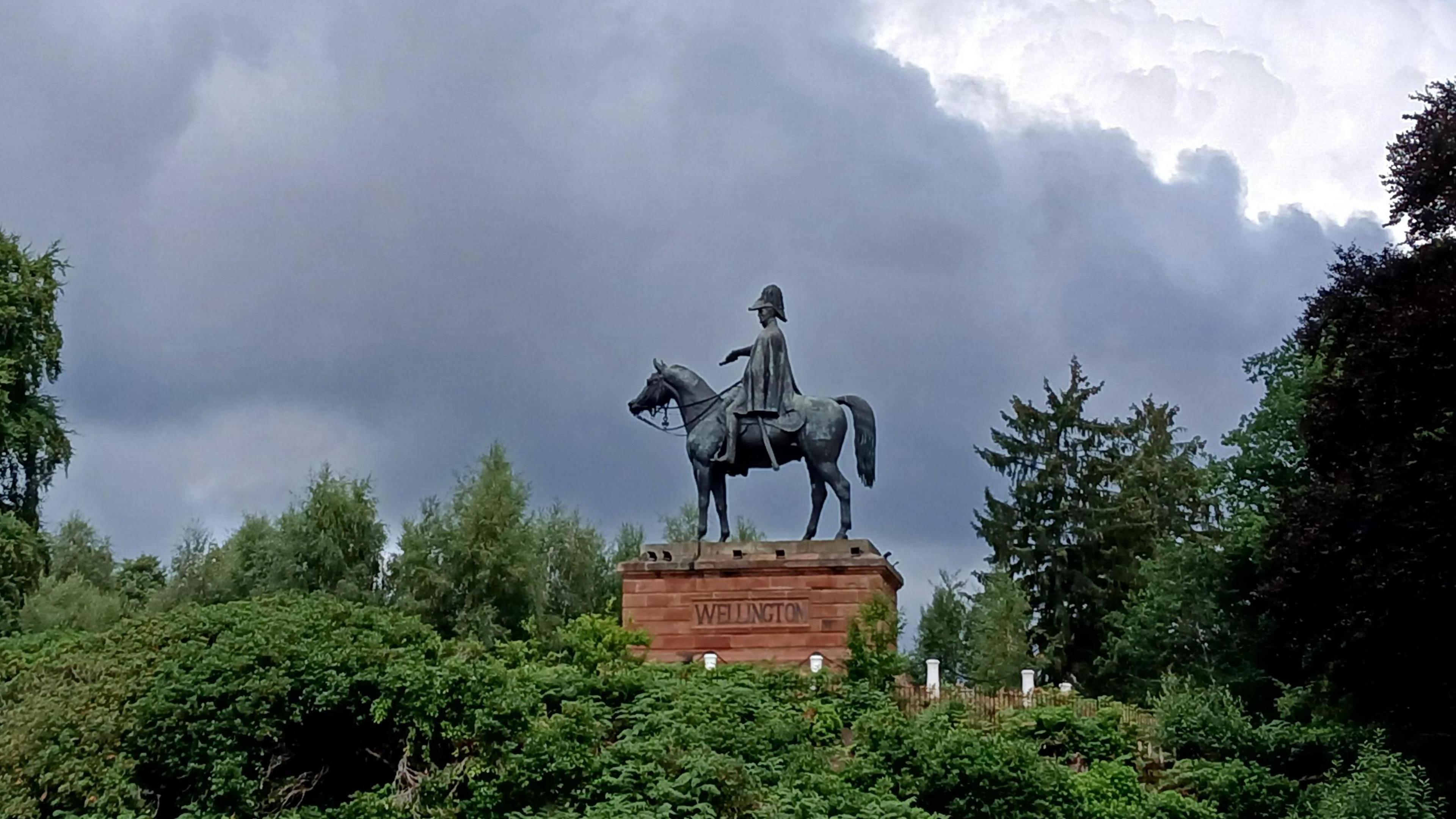 A statue of the Duke of Wellington on horseback sits on a red brick plinth surrounded by trees. Dark clouds are building in the sky
