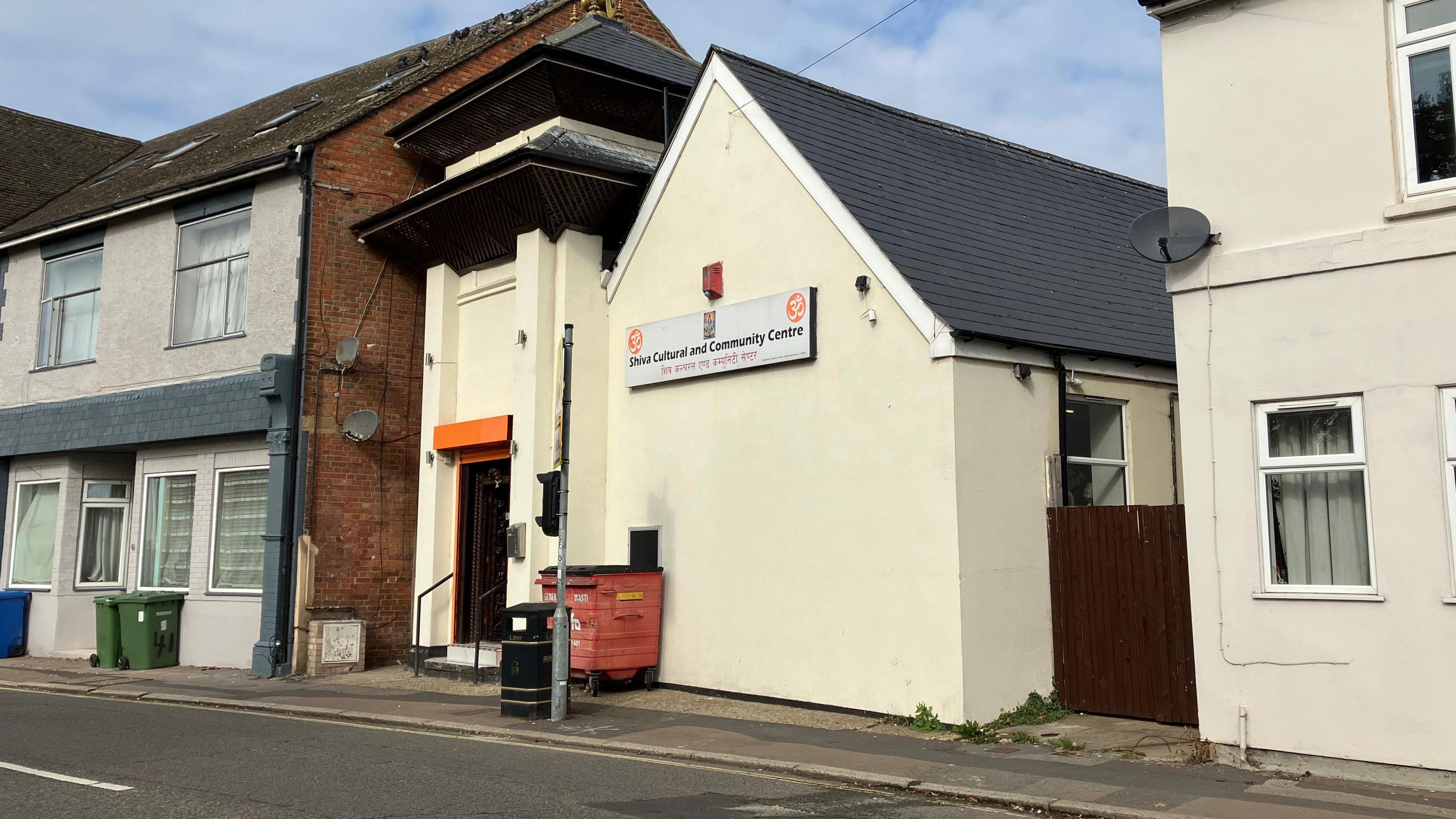 The outside of the Shiva Cultural and Community Centre, which is a low building in cream render. It opens directly on to the pavement, and there is a house next door. A large red wheelie bin is on the pavement outside the centre.