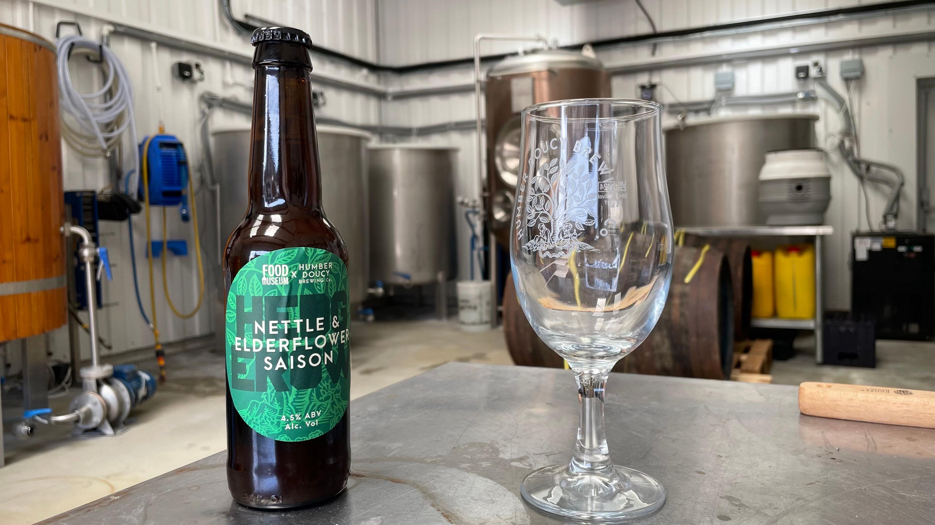 A bottle of Humber Doucy beer and an empty half pint glass, which also has the brewery's name and logo on it. Both are sitting on a metal table in the brewery's warehouse. Brewing machinery can be seen in the background.