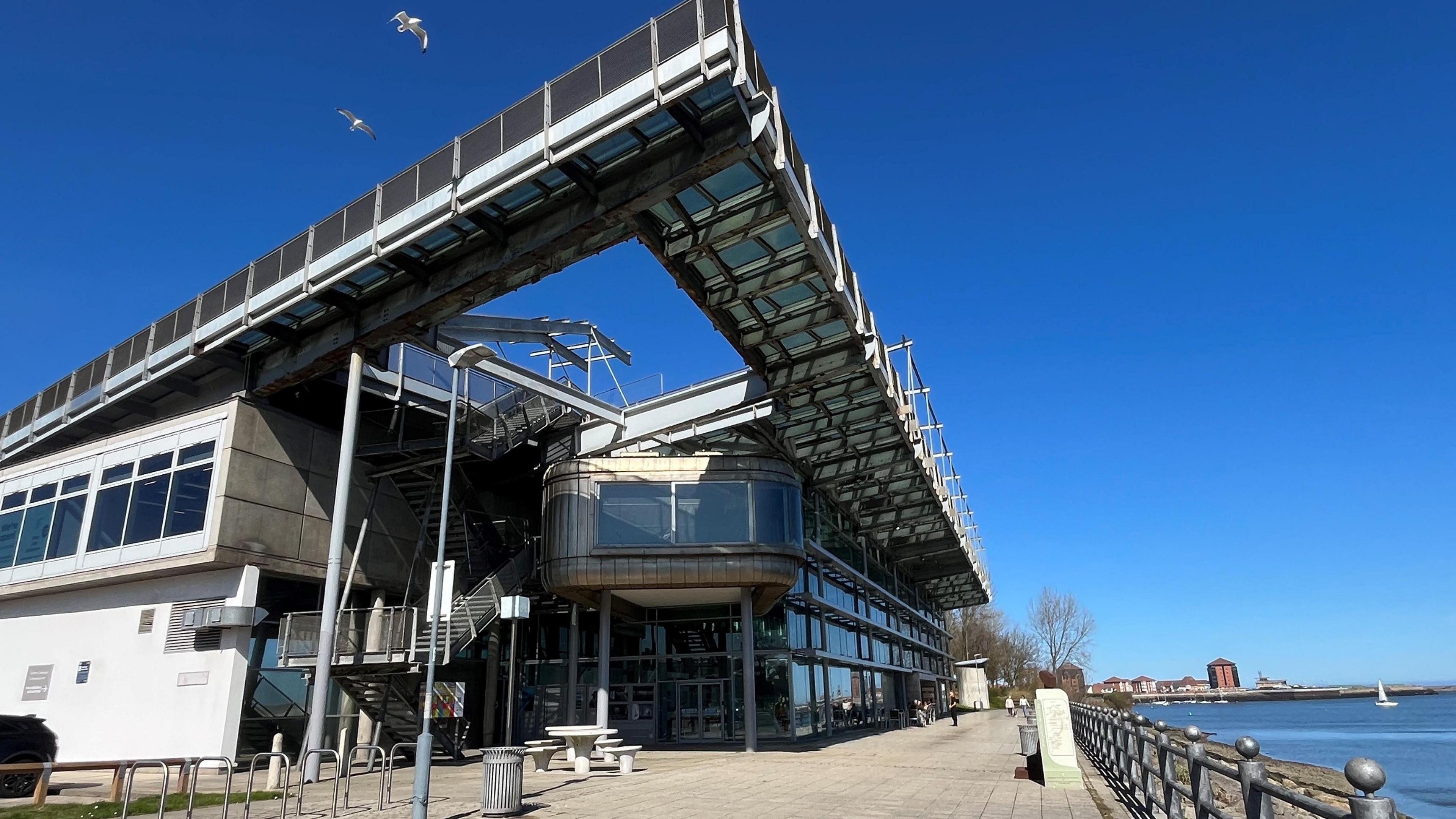 The National Glass Centre is a tall building made of metal panels and glass, overlooking the river Wear. There is seating space and a cafe in front of it, with a dog walker in the distance. Two seagulls are flying above the building.