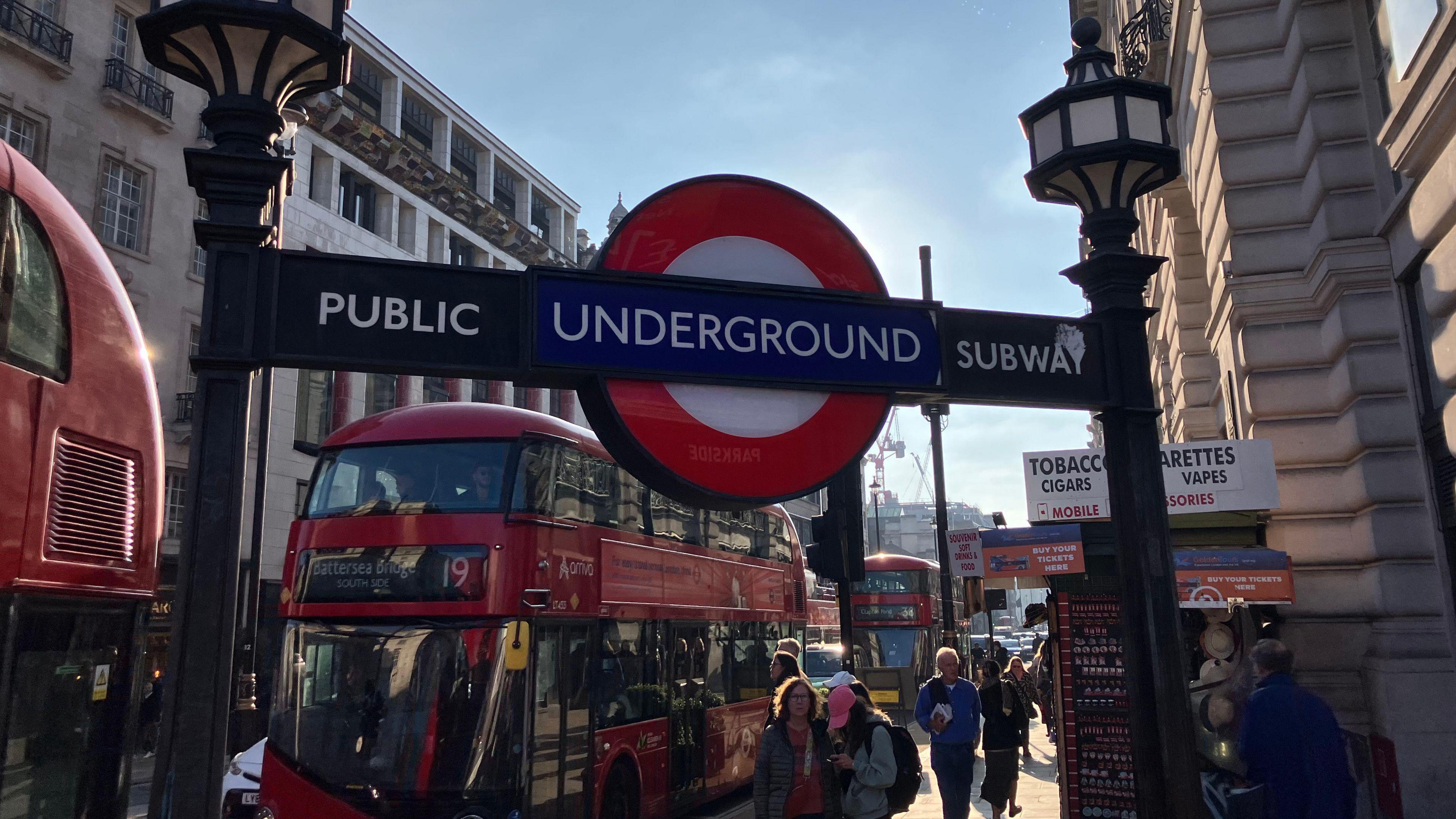 In the foreground is a blue and red Tube roundel between 'Public subway' sign. In the background is a red double decker 19 bus and people walking along the pavement