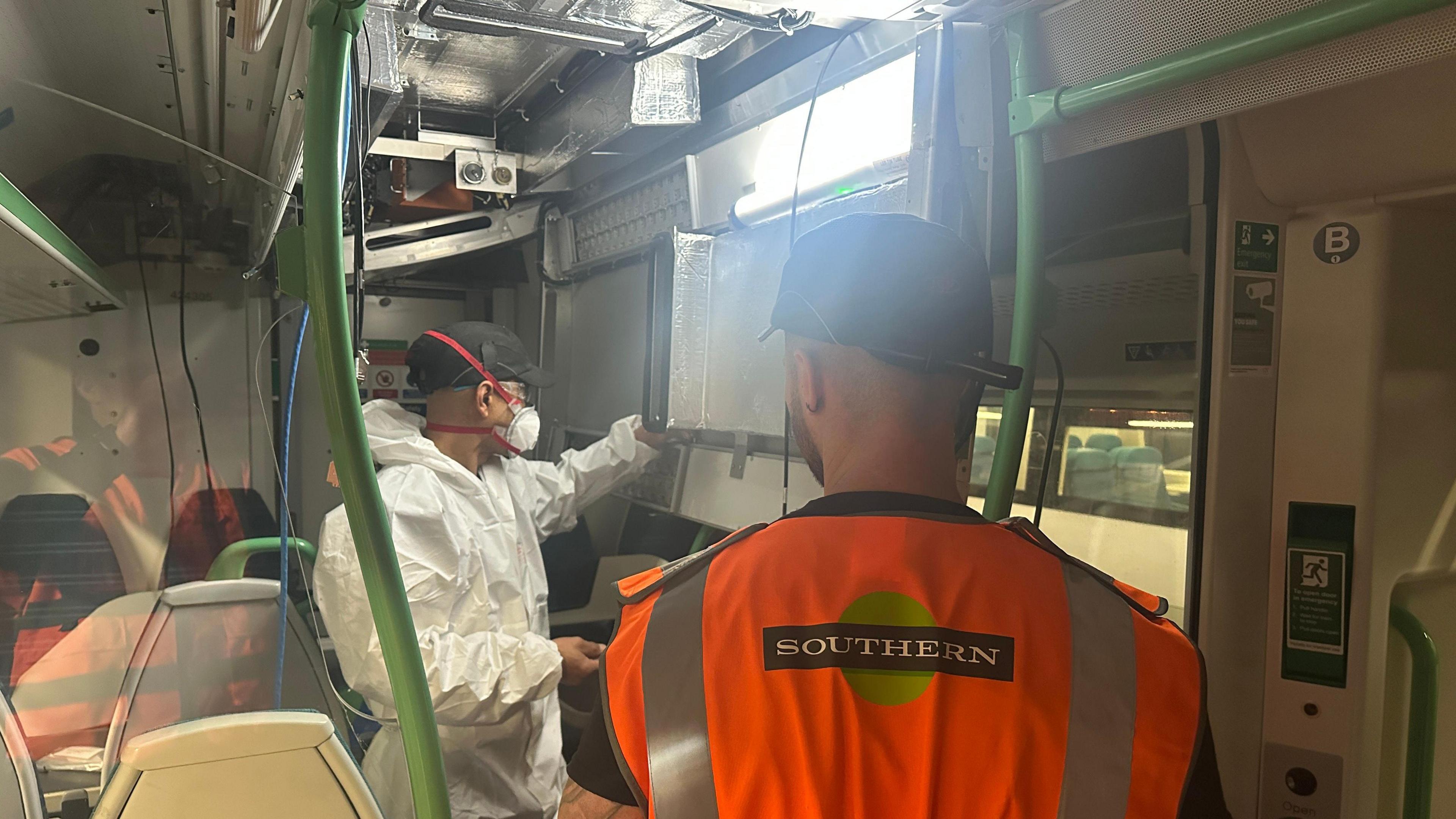 Two railway maintenance workers stand inside a train carriage - one in a Southern-branded orange jacket, the other in a white protective suit and face mask