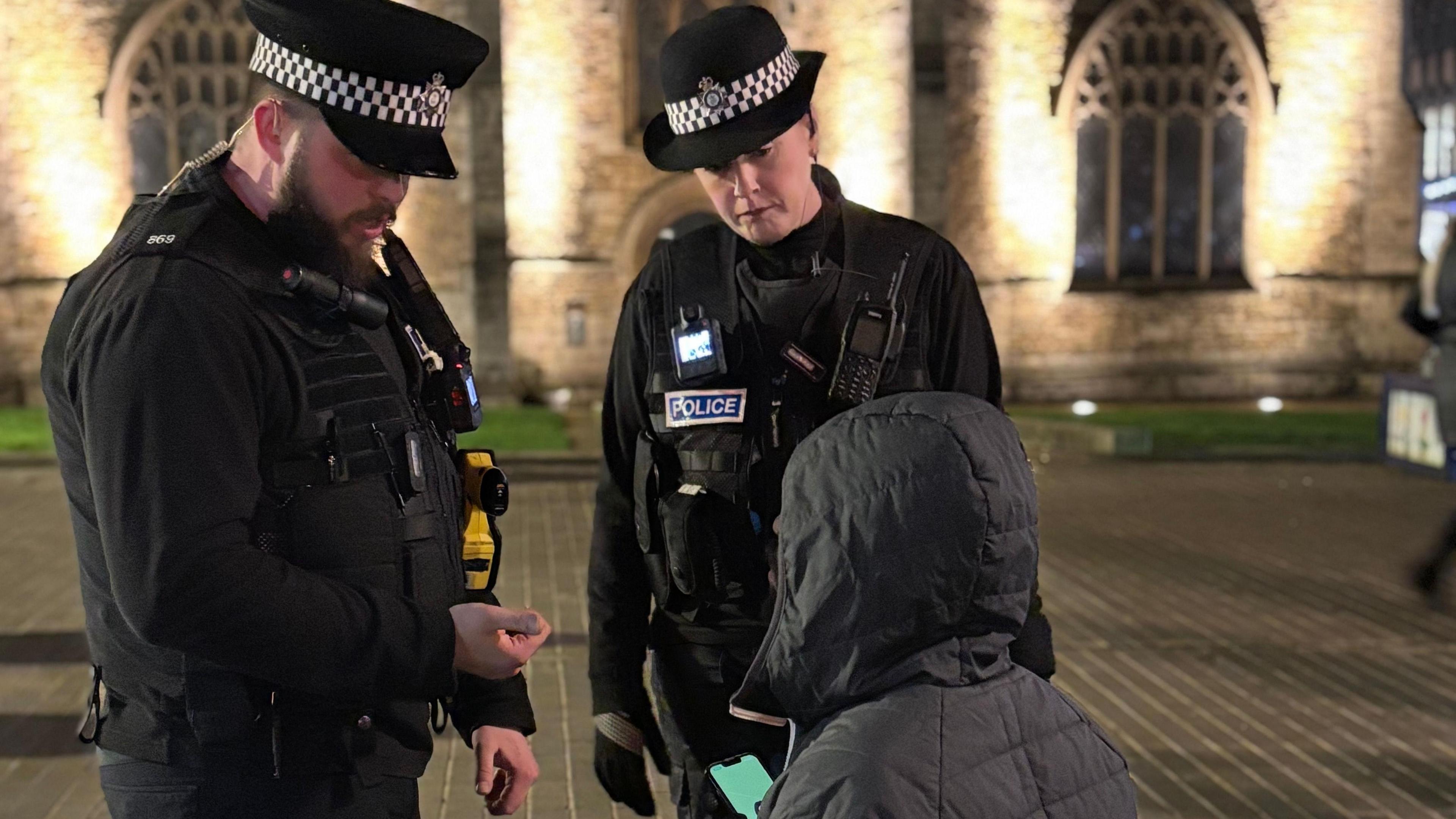 Police officers in black uniforms- speaking to a person wearing a grey hood and jacket- with the back facing the camera 