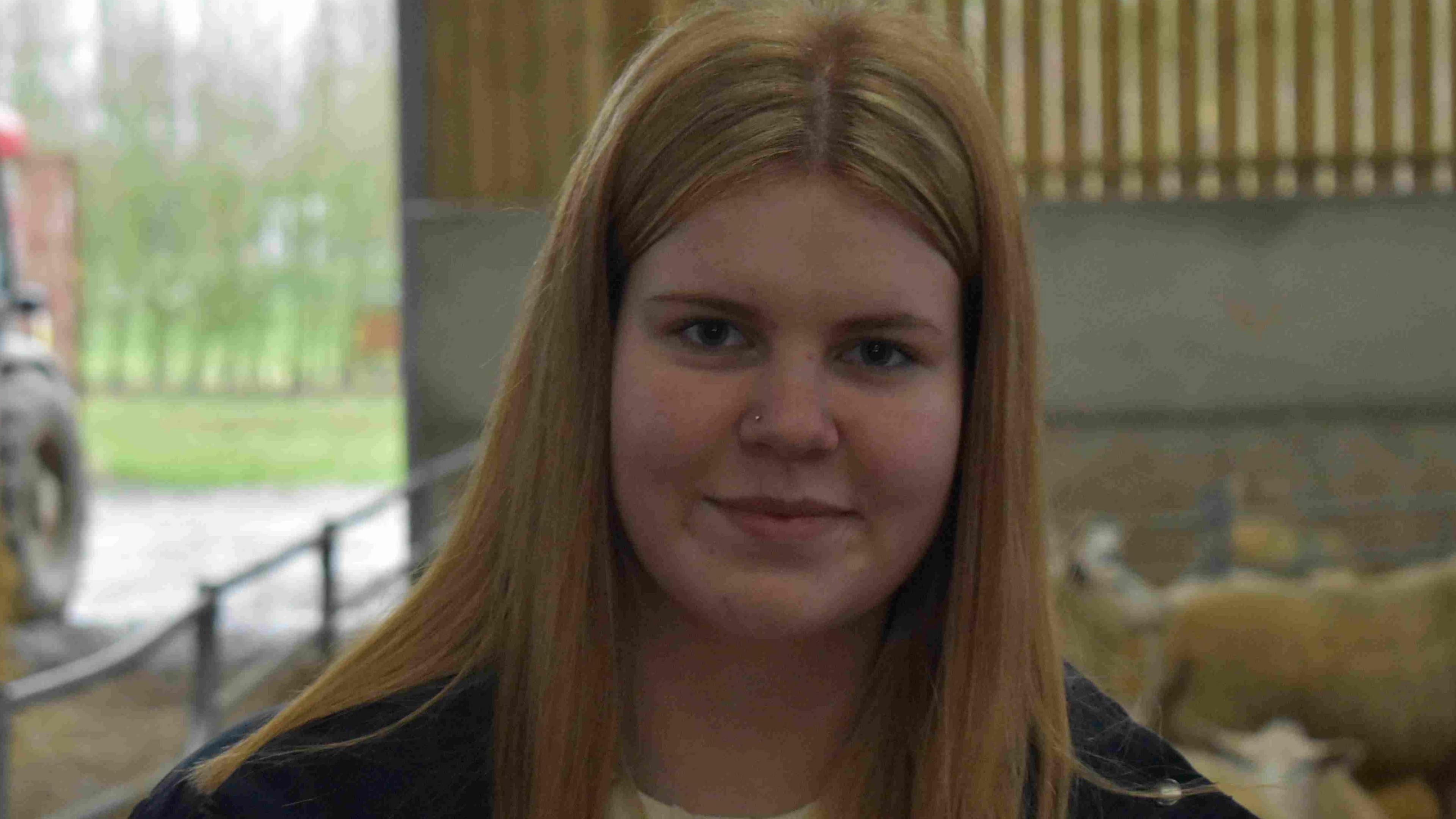 Daisy has long blonde hair. She is staring at the camera while standing in a barn in front of a sheep pen. To the left of the image a tractor can be seen driving into the barn.