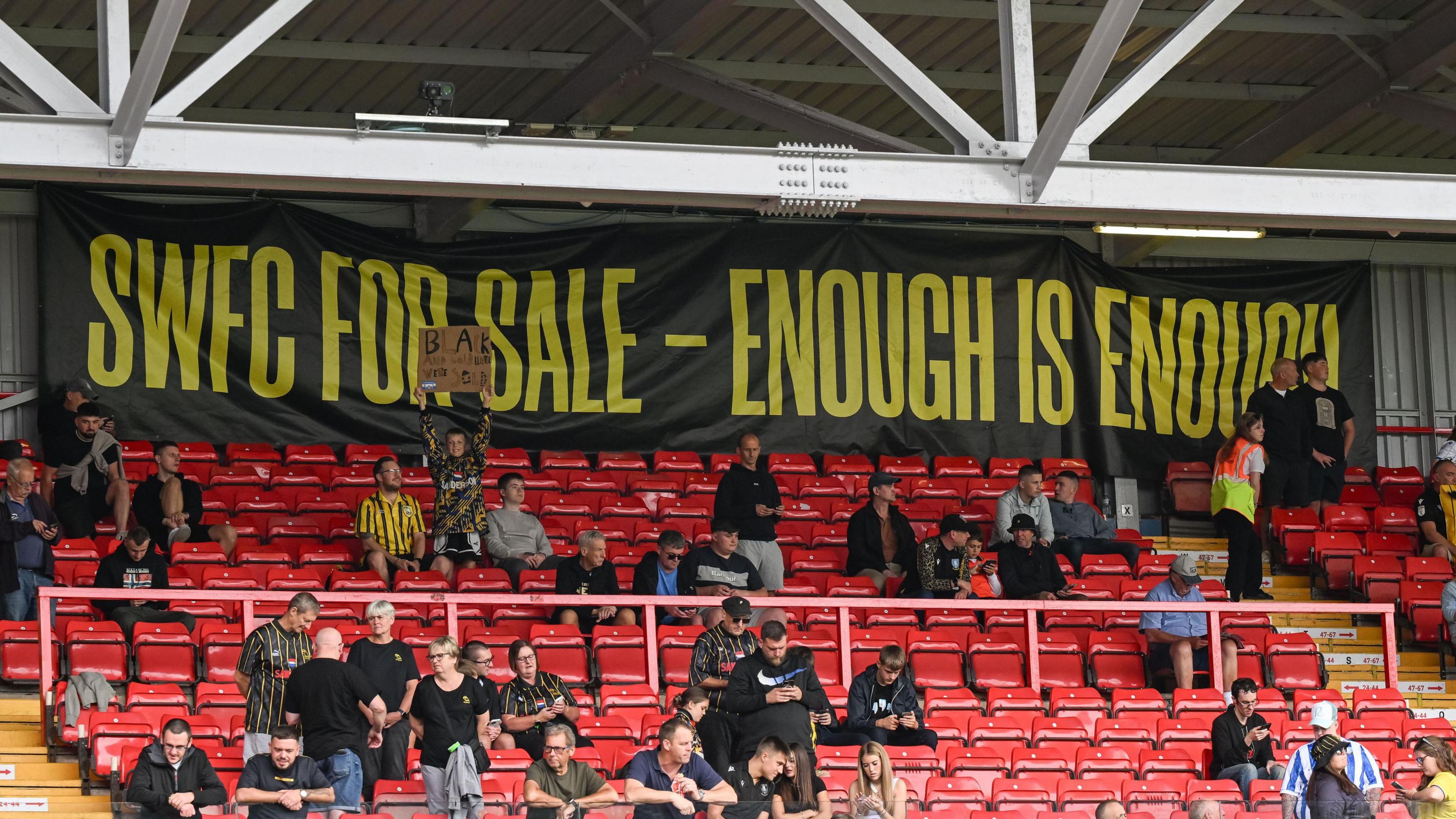 Black banner that says 'SWFC for sale - enough is enough' at the back of a stadium with red seats where numerous people sit