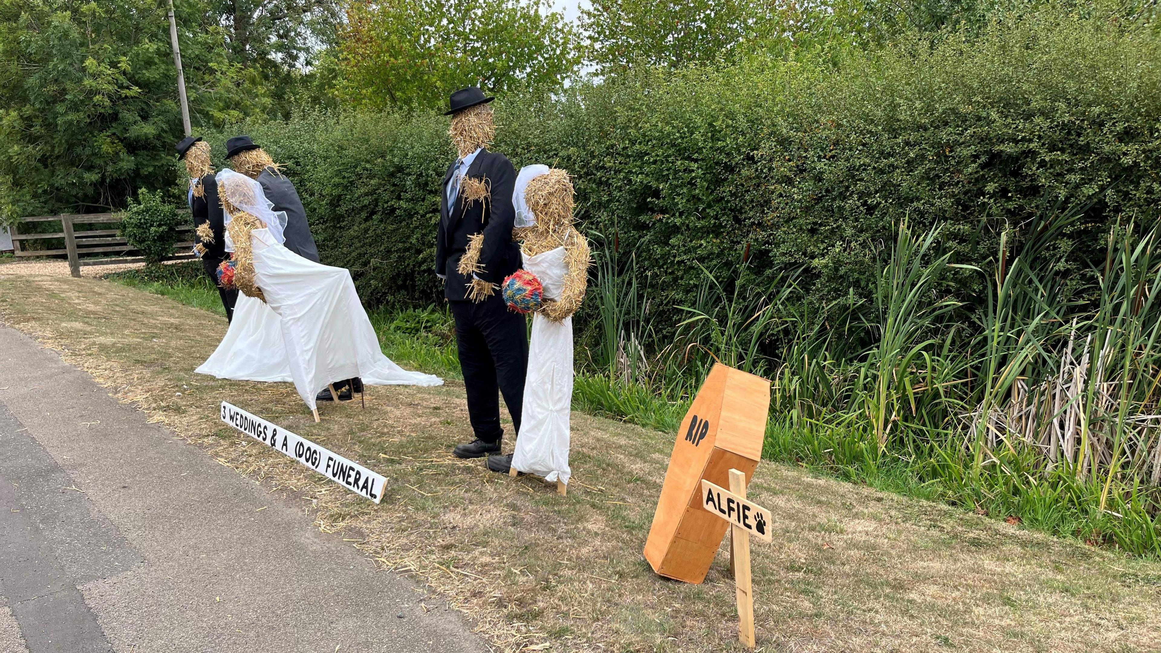 Six life-size figures of three couples in wedding dresses and suits are standing on a grass verge outside a house on the High Street in a rural location, a small wooden coffin with RIP written on the lid next to a sign which says 'Alfie' on it.
A longer white sign stands in front of the display and says 'three weddings and a dog funeral'. Behind the scarecrows is a tall 2.5m (8ft) hedge and a gravel driveway beyond that.