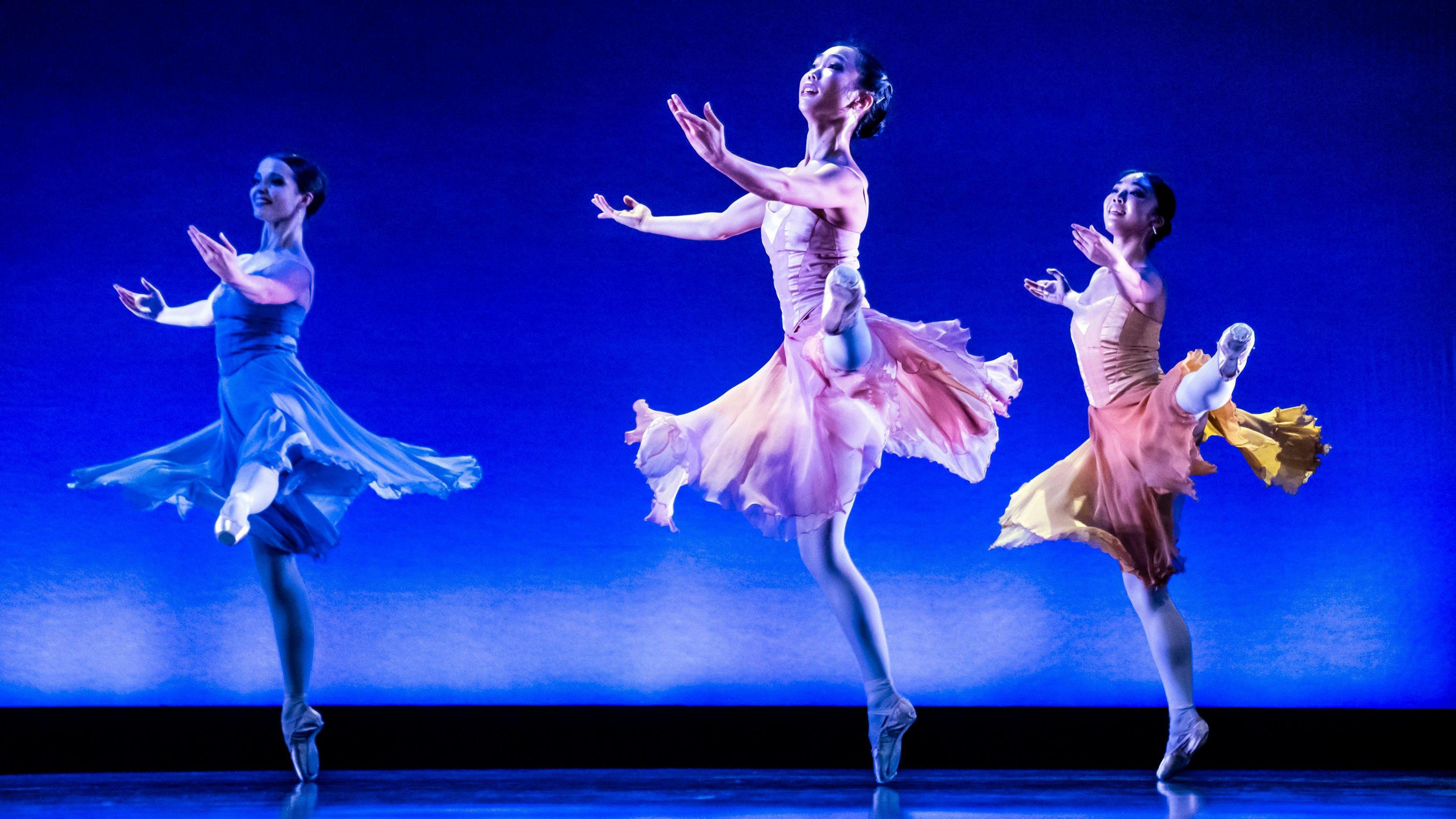 Three female dancers during a dress rehearsal at the Northern Ballet's Stanley and Audrey Burton Theatre in Leeds, West Yorkshire. The dancer in the middle is wearing a pink tutu, the one on the left is in blue and another  on the right is in orange and yellow. They are all wearing white tights and are dancing against a blue background.