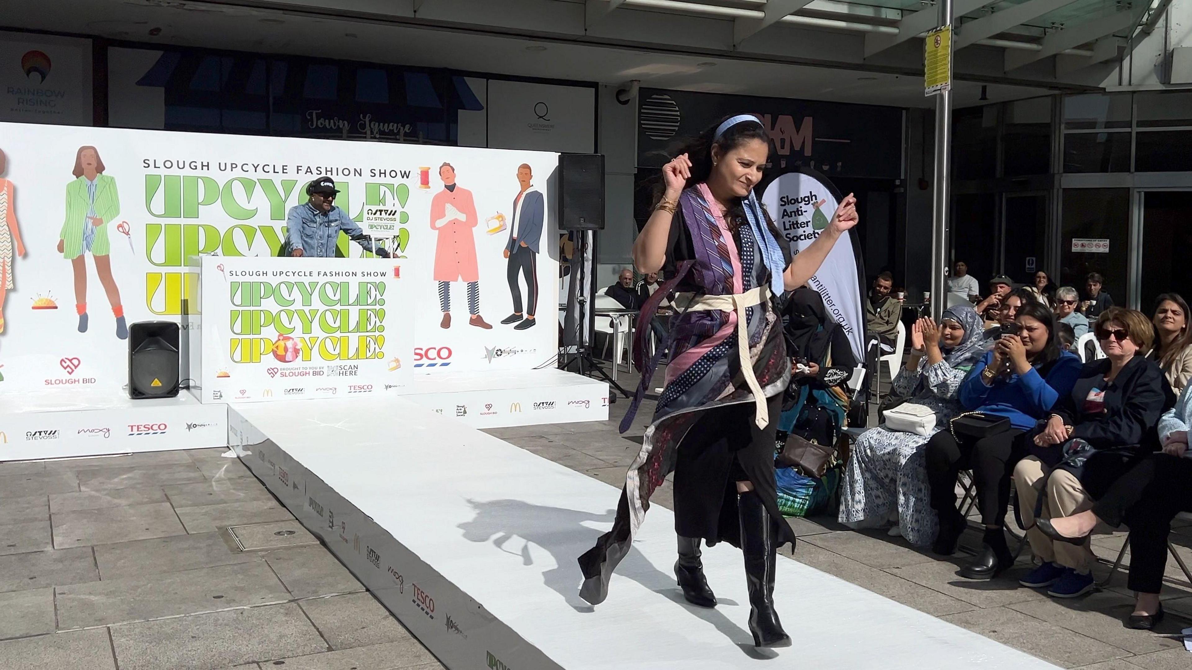 Quesia Rahim strides down the catwalk as people look on. Her dress is shaped out of different coloured ties. She has long dark hair and a headband, which might also be a tie!