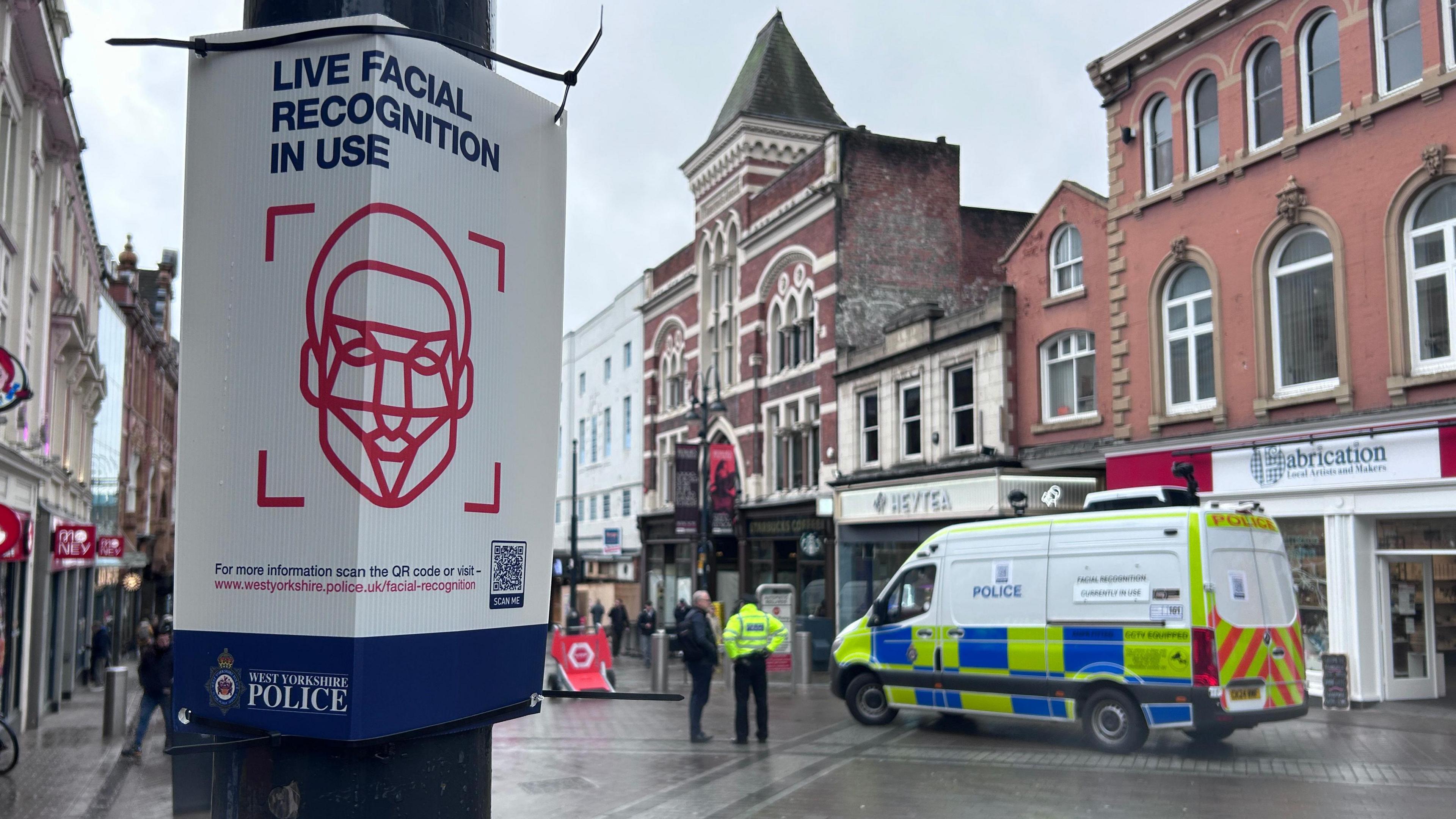 Briggate, a main shopping street in Leeds. There is a close-up of a sign on the lefthand side of the shot, saying that live facial recognition is in use. On the other side of the street, a police van can be seen. There is a police officer standing next to it. This part of the street looks quiet.