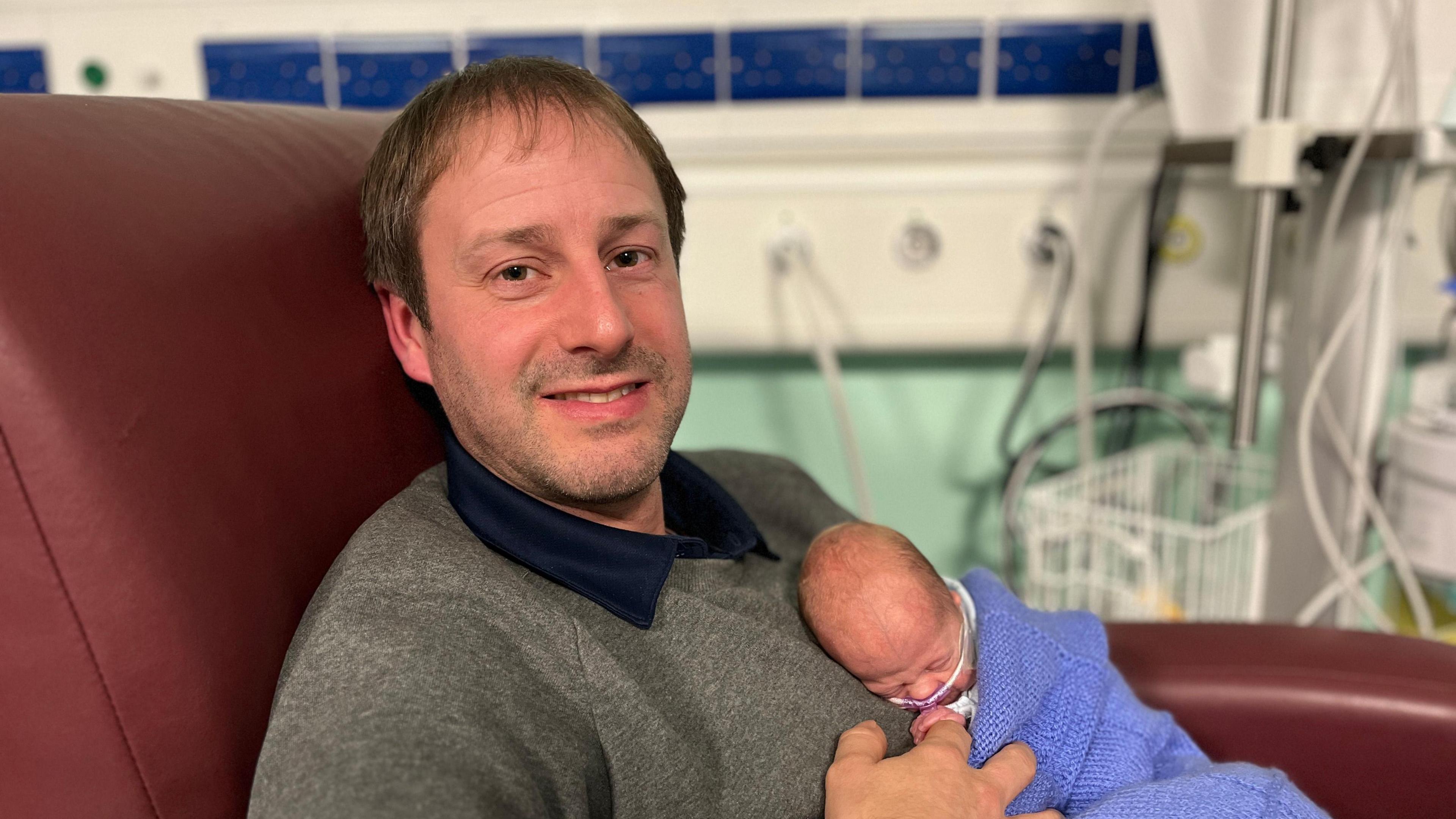 Tony has short brown hair and is wearing a navy shirt and grey jumper. He is sitting in a red hospital chair holding baby Elliot, who is wrapped in a blue blanked, on his chest. Elliot has a feeding tube.