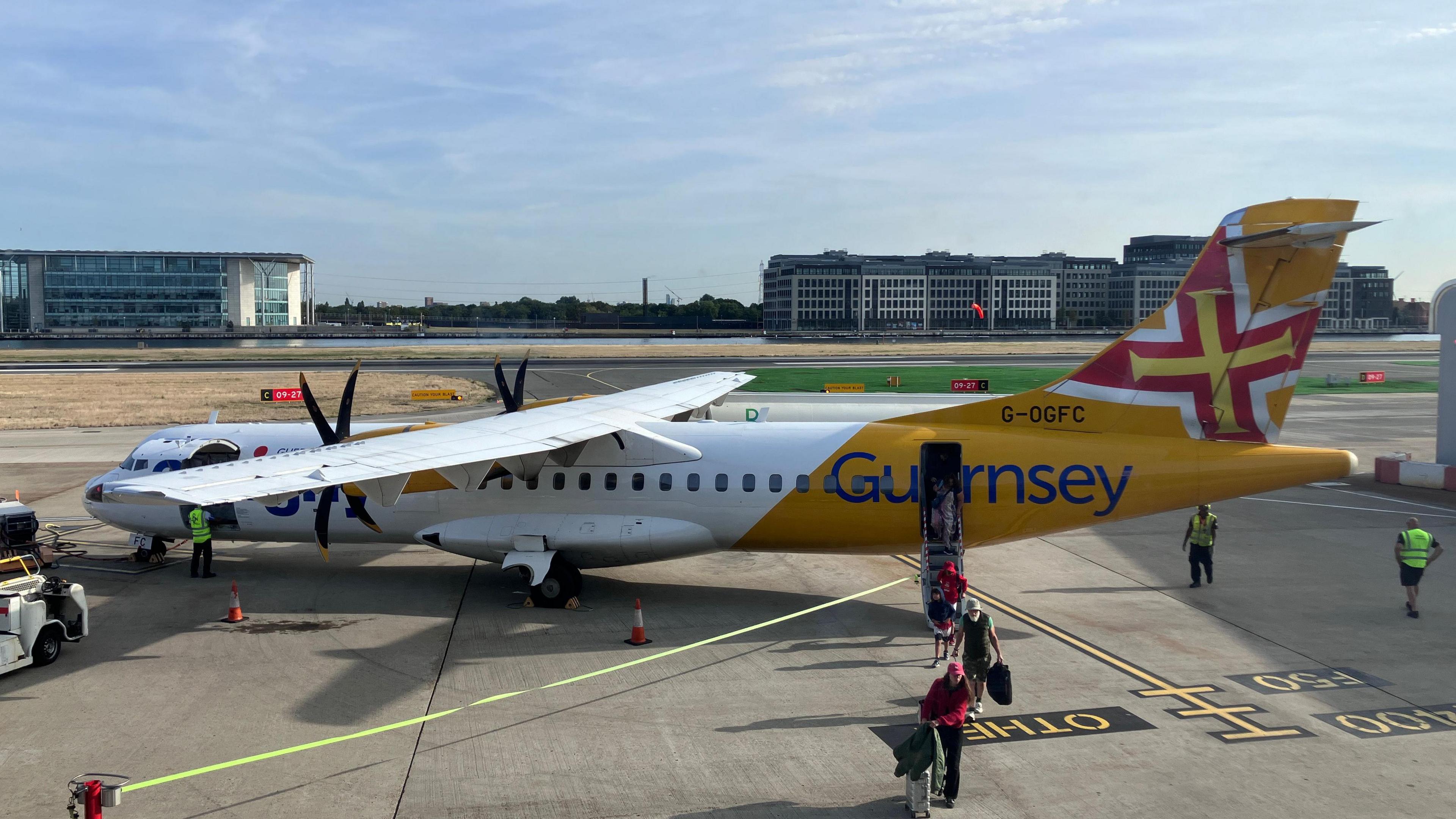 A white and yellow propellor aircraft with the word Guernsey on the side and a Guernsey flag on the tail. The ramp is down and passengers are disembarking. 