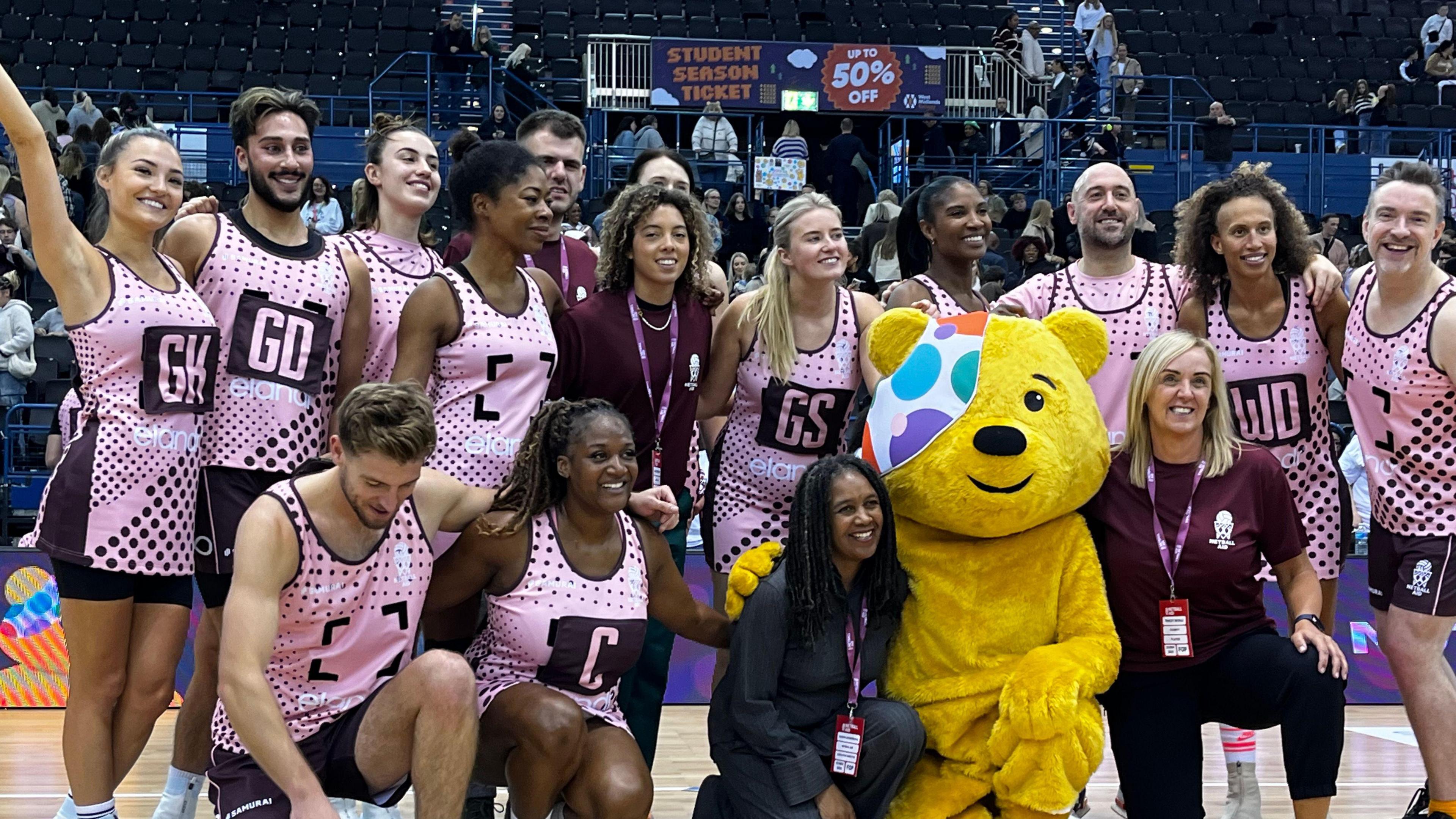 A group of people in pink netball kits stand on a netball court and smile. Someone dressed in a Pudsey bear suit kneels in front of them.