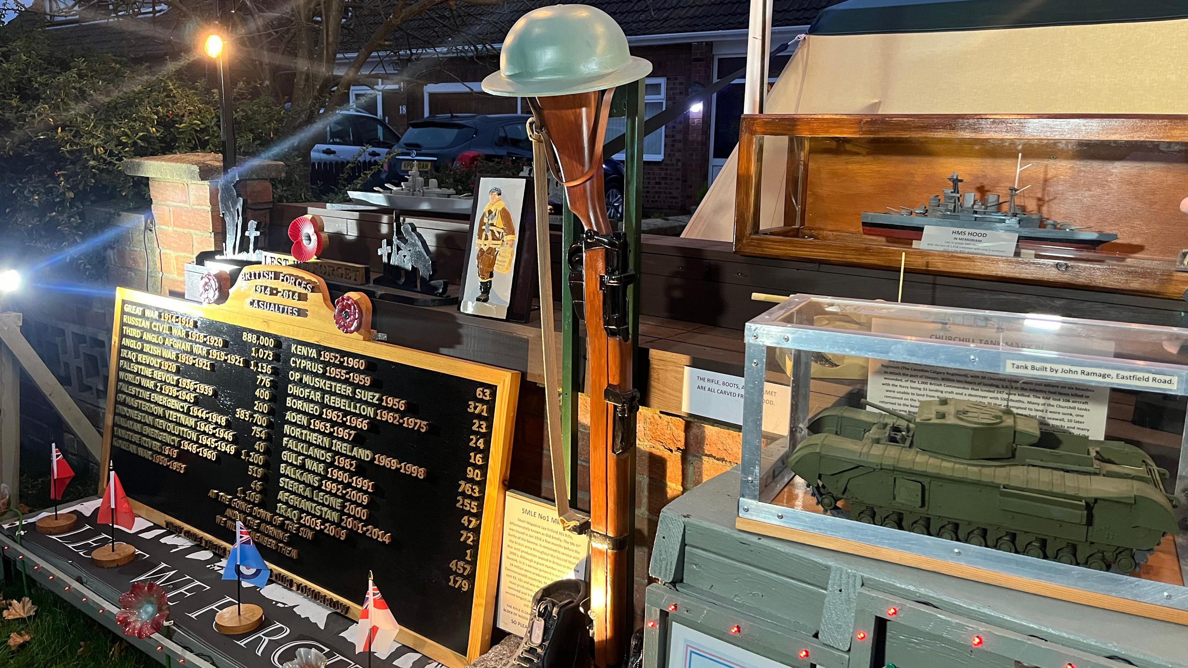 A photo of the Remembrance display. There is a board with all of the wars listed. There is a hand-carved rifle with a war helmet on top and a boot at the bottom. 