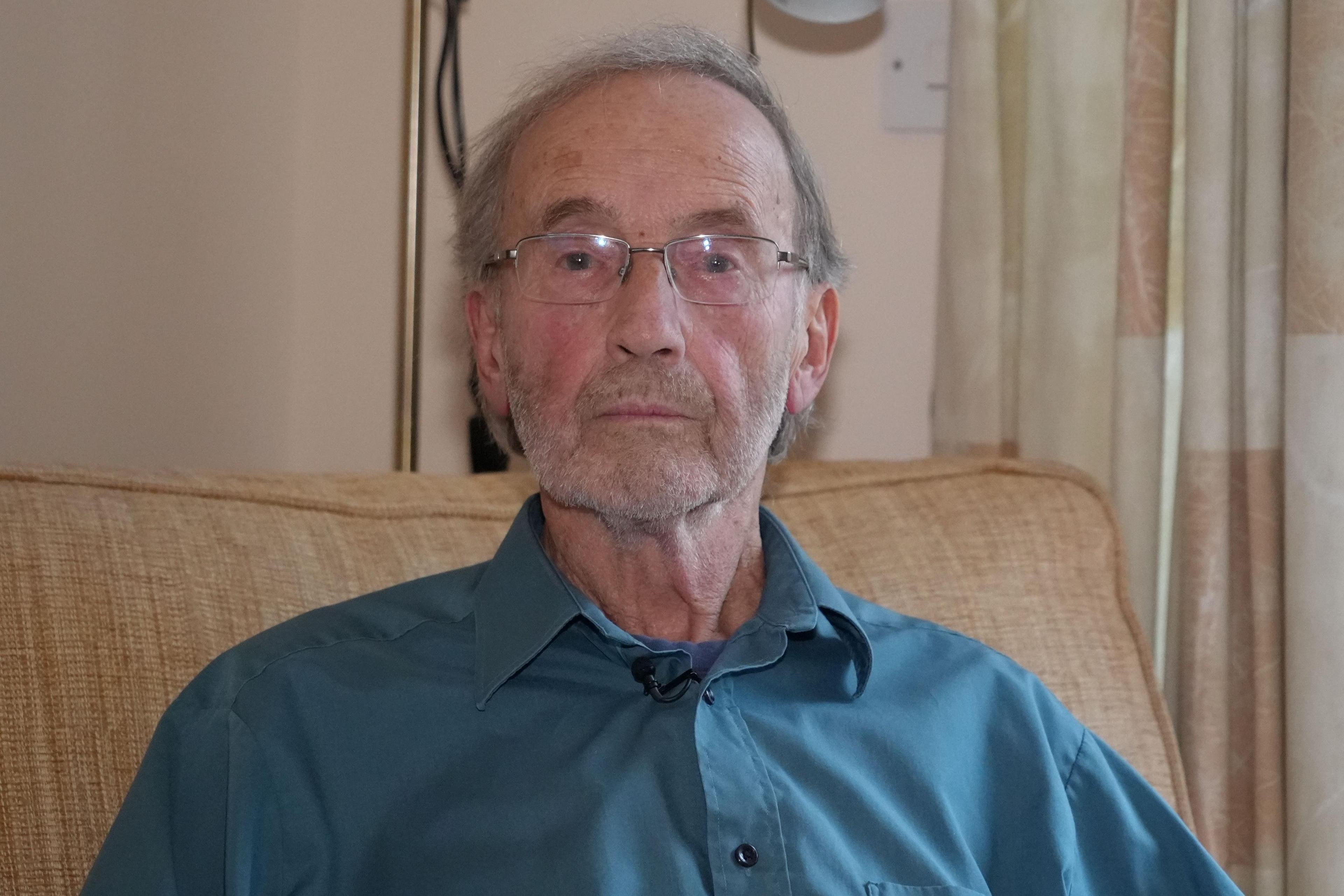 David Tate sitting on a sofa with a blue shirt, wearing clear glasses. His hair is greying. He is looking at the camera lens.