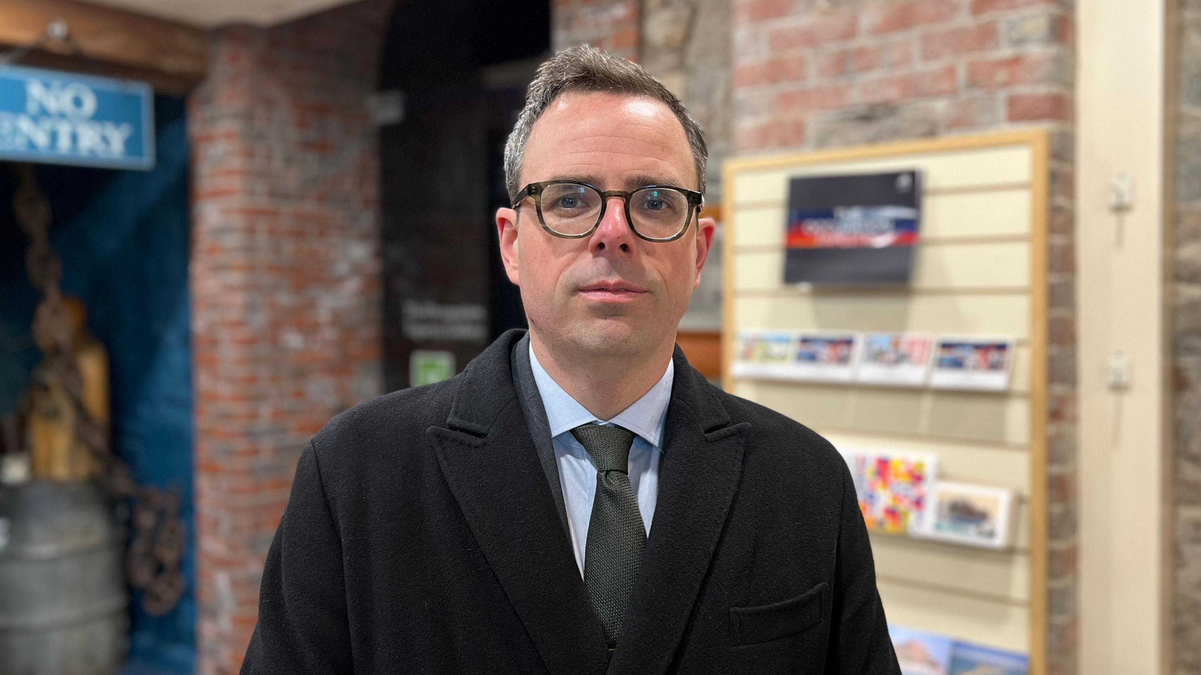 A man wearing a black coat over a suit with a blue shirt and grey tie, wearing black glasses standing in a museum - the background is blurred.