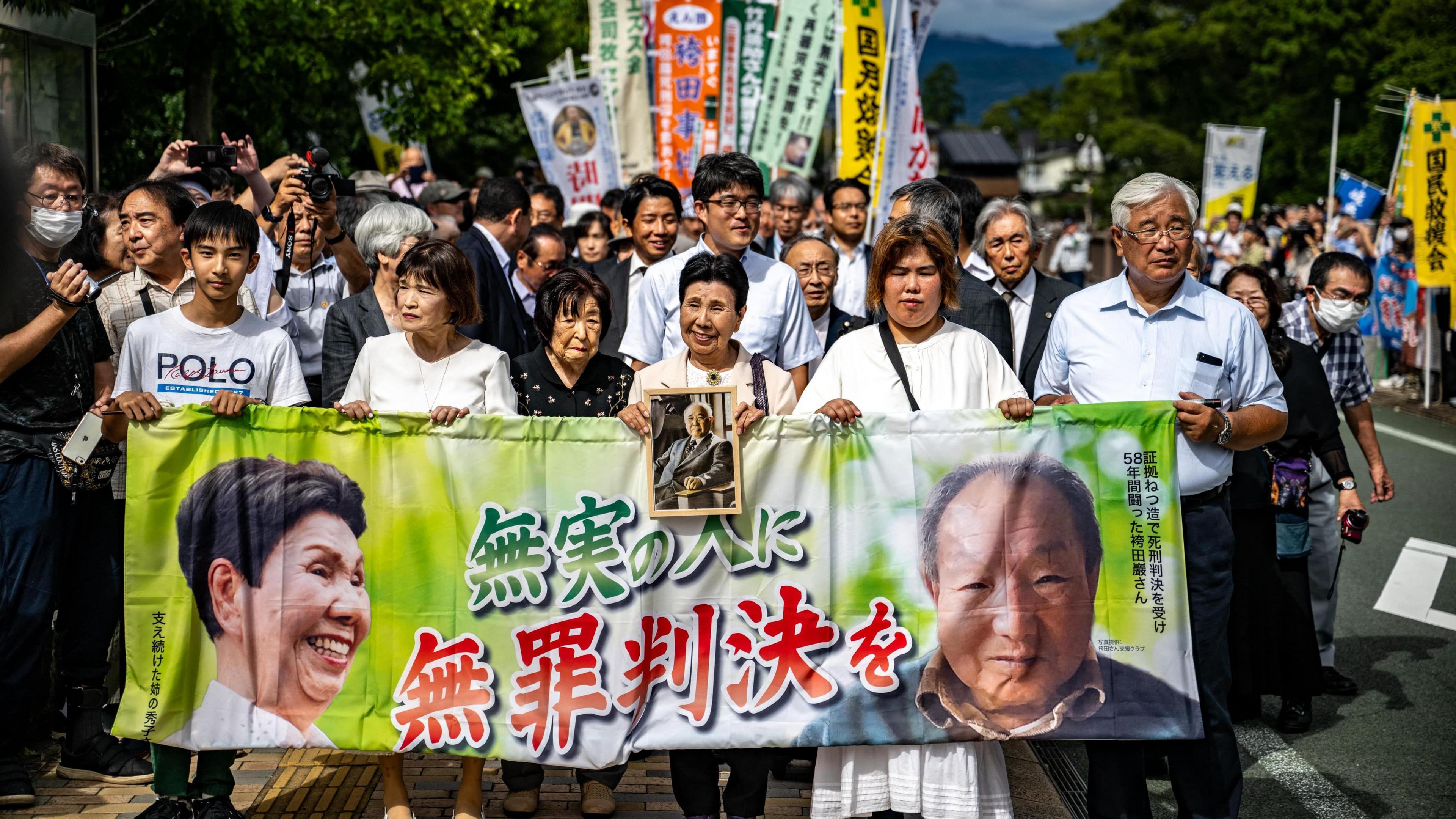 A crowd of people walks down the street holding a banner showing the faces of Iwao Hakamata and Hideko Hakamata 