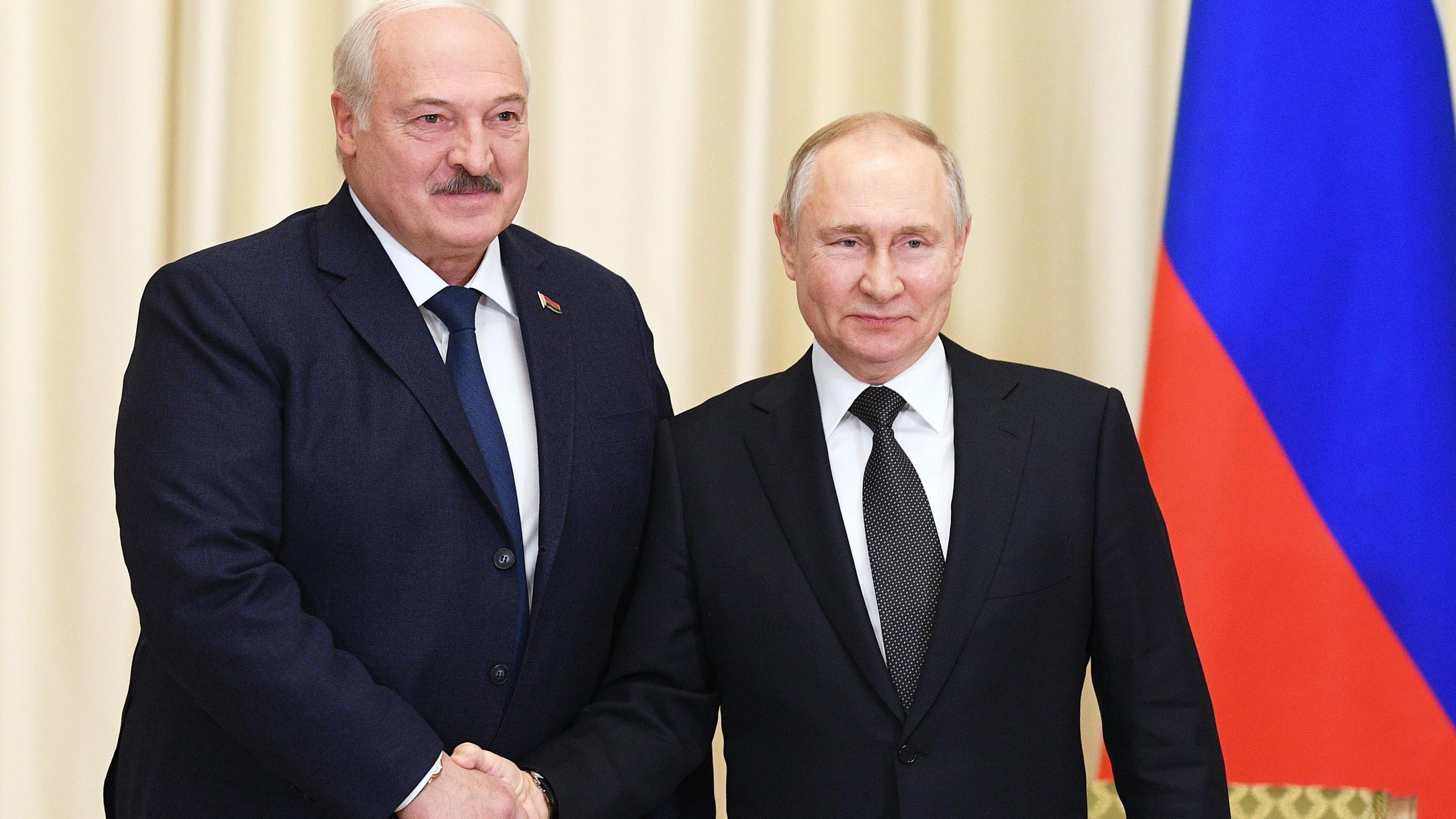 President of Belarus Alexander Lukashenko, wearing a dark suit, white shirt and dark blue tie, shakes hands with Russian President Vladamir Putin who is waring a dark suit, white shirt and black tie. A Russian flag can be seen to the right of the shot.