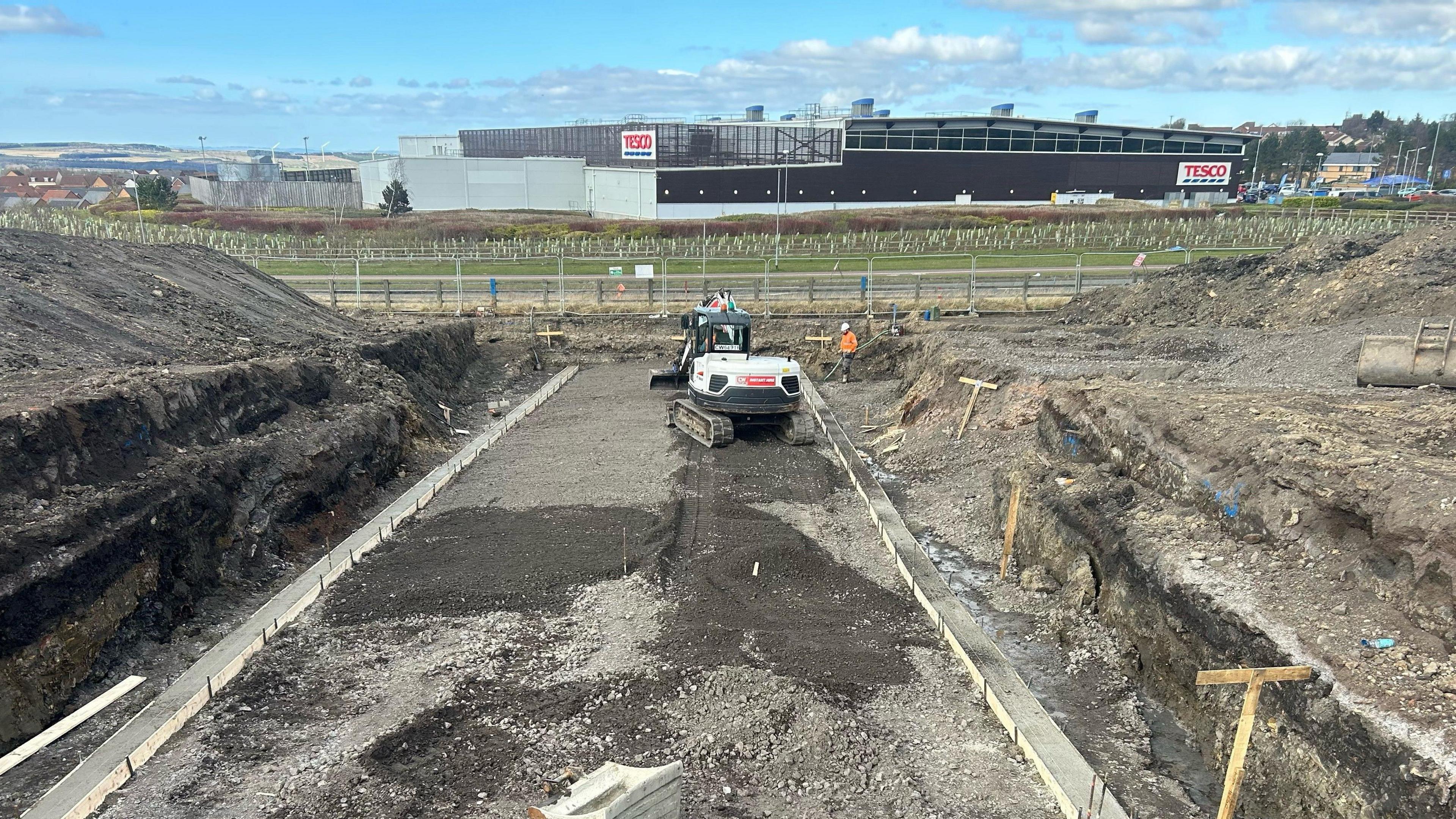 A bulldozer at the site of the Shotley Bridge Hospital.  A section of the land has been dug out. There is a Tesco store behind the building site. 