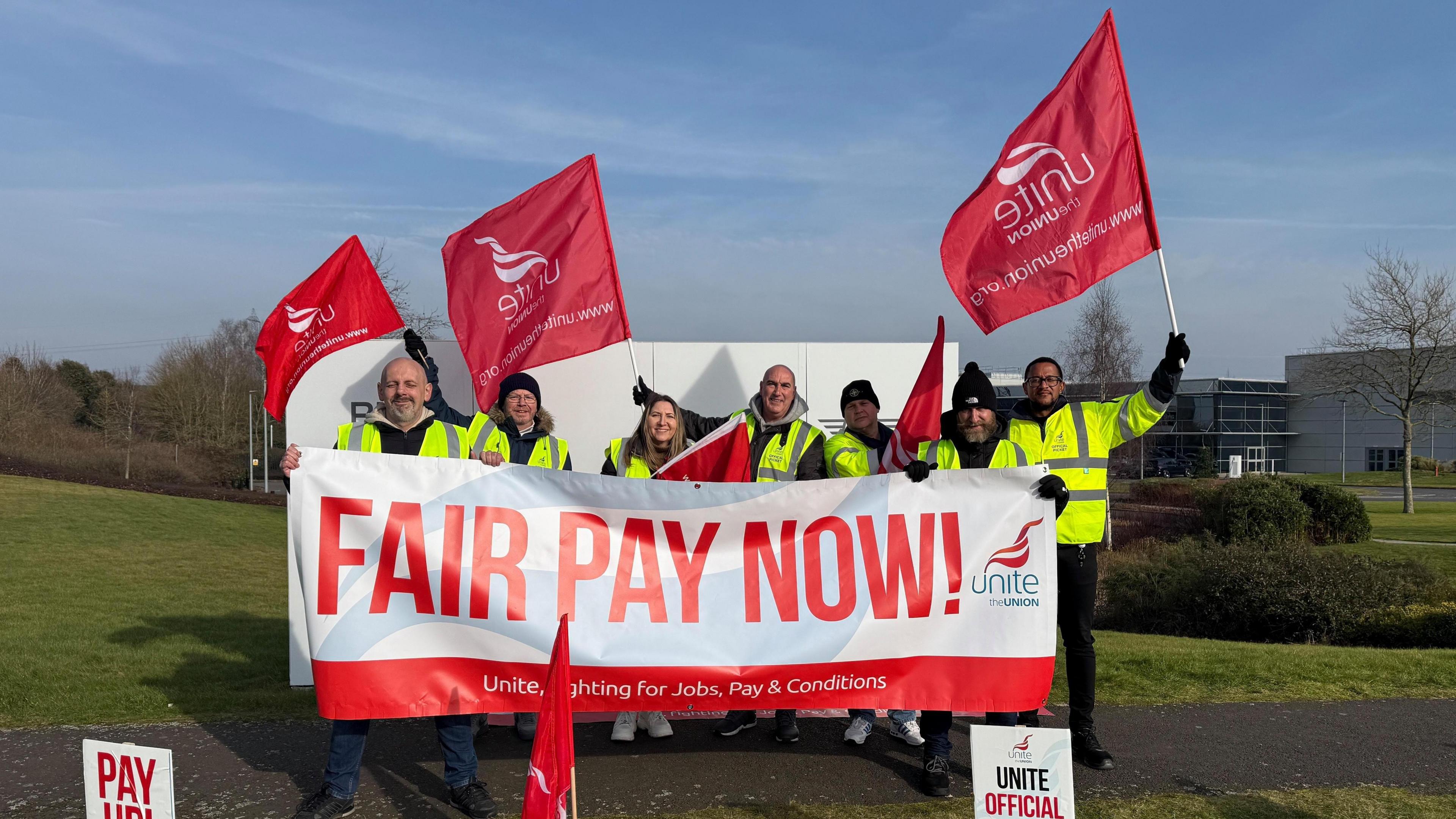 A group of seven people in a hi-vis holding signs saying 'fair pay now'