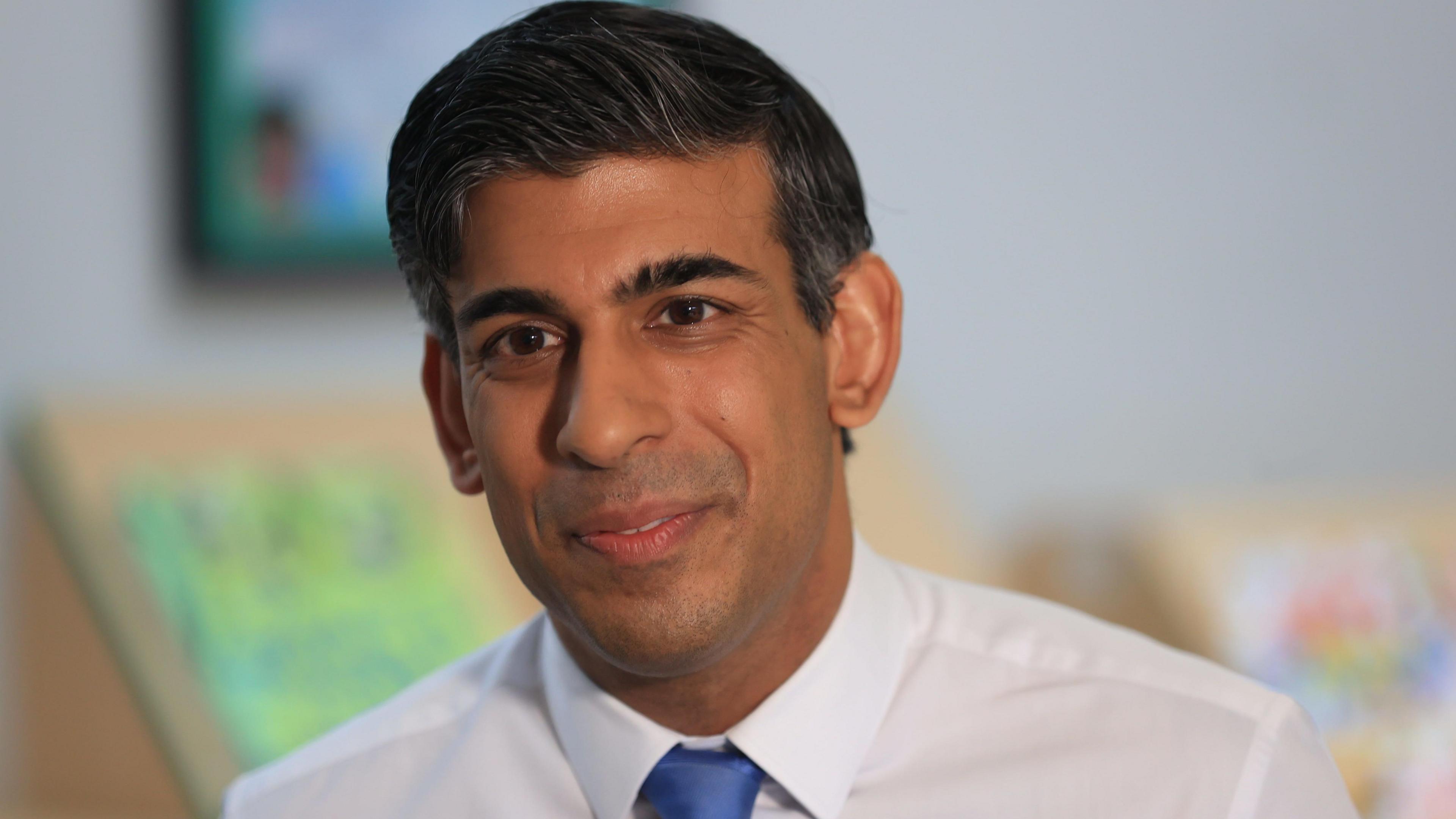 Rishi Sunak is smiling and wearing a white shirt and a blue tie