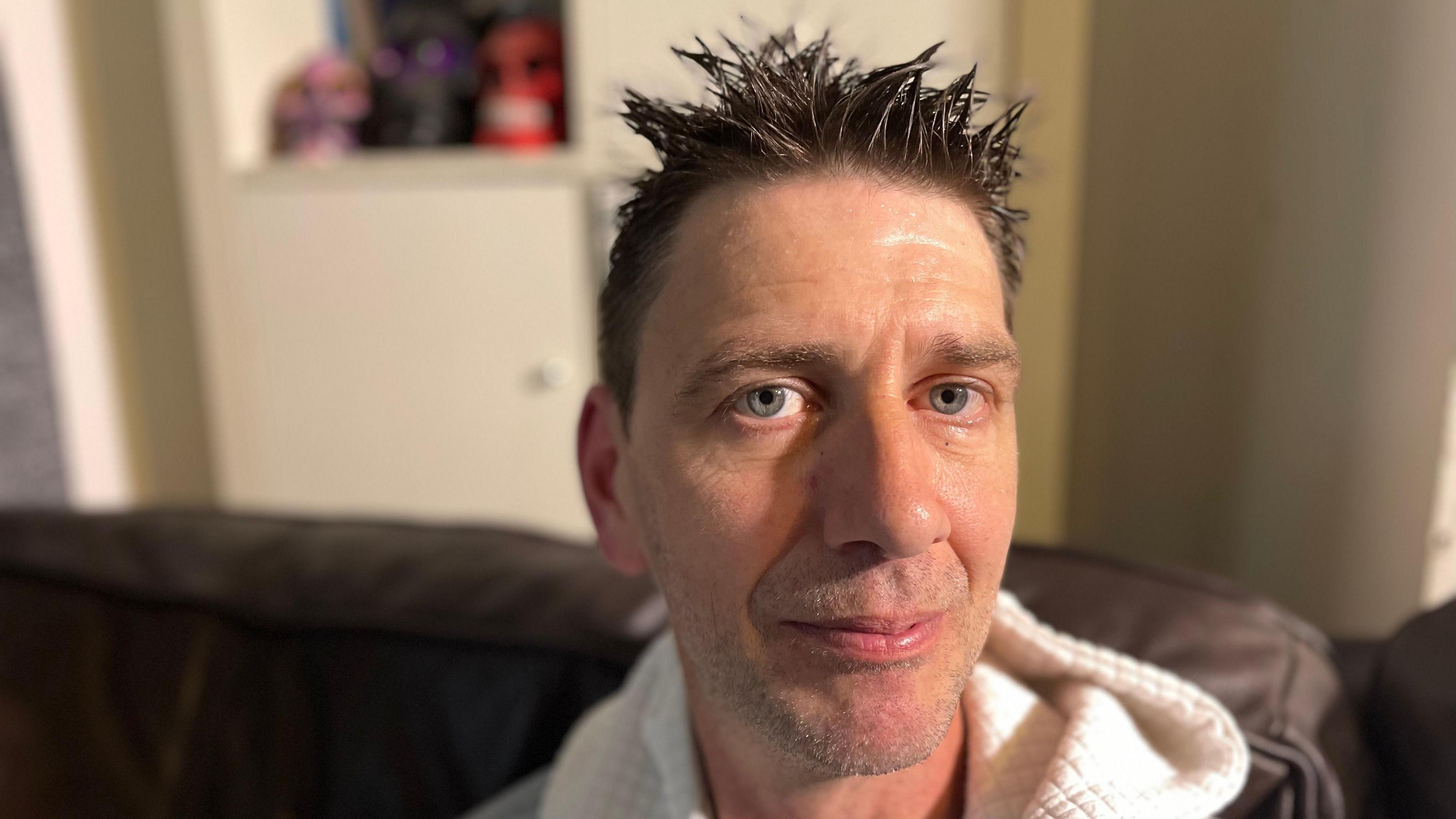 Man with spiky hair wearing grey and white hoodie sitting on a brown leather sofa.