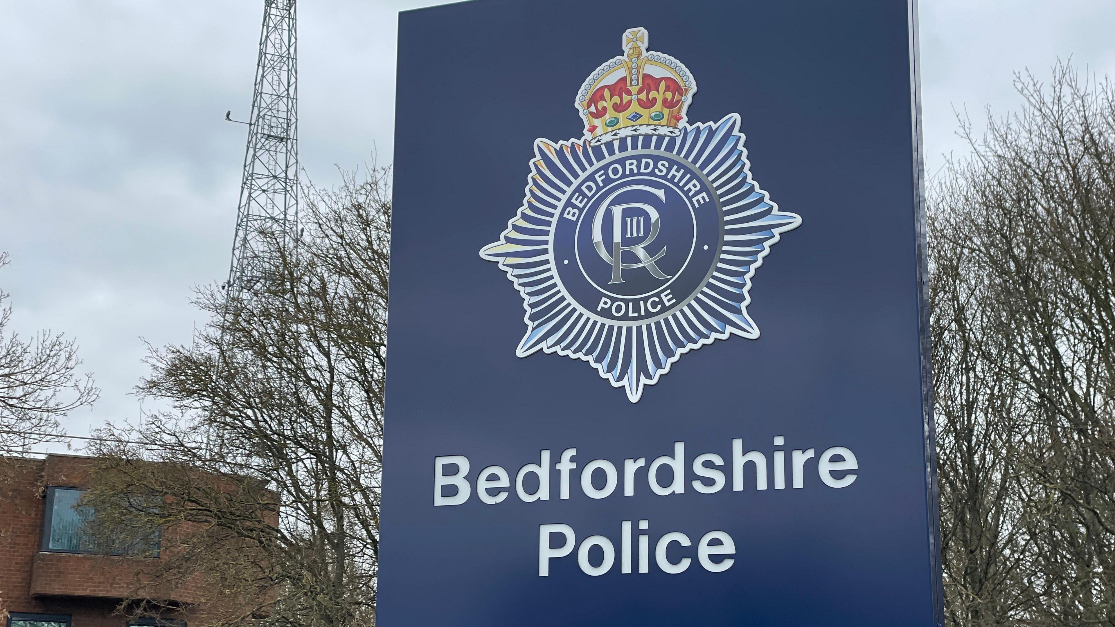 A rectangular blue sign with the Bedfordshire Police crest featuring a golden crown on top of a seven-sided star shape. King Charles' insignia, a C and an R and a Roman numeral 3, are inside a circle inside the shape. The words "Bedfordshire Police" arc around the circle in capital letters. 