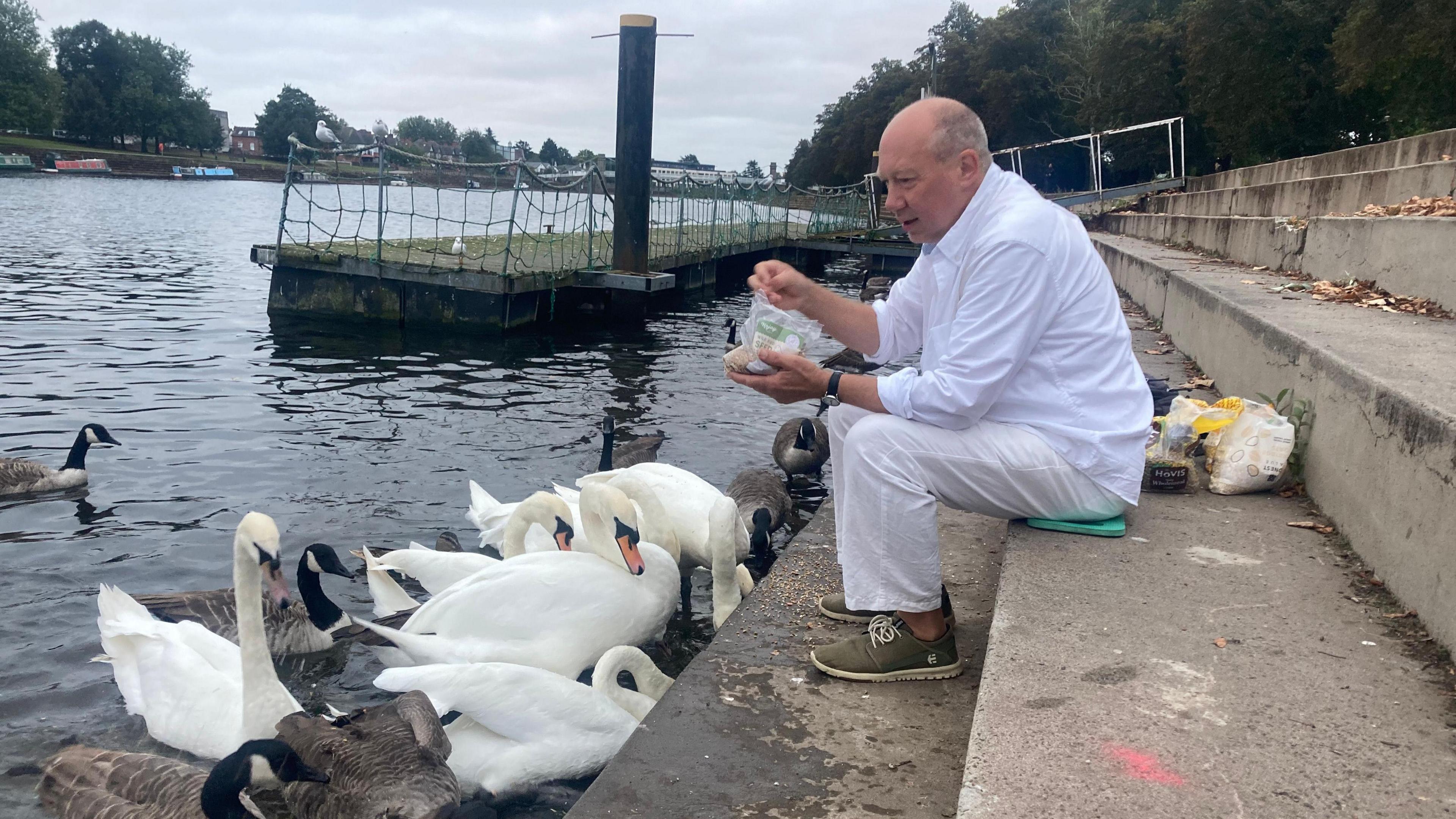 Simon Withers, wearing white trousers and shirt, sits on concrete steps by the River Trent in Nottingham feeding a bevy of swans.