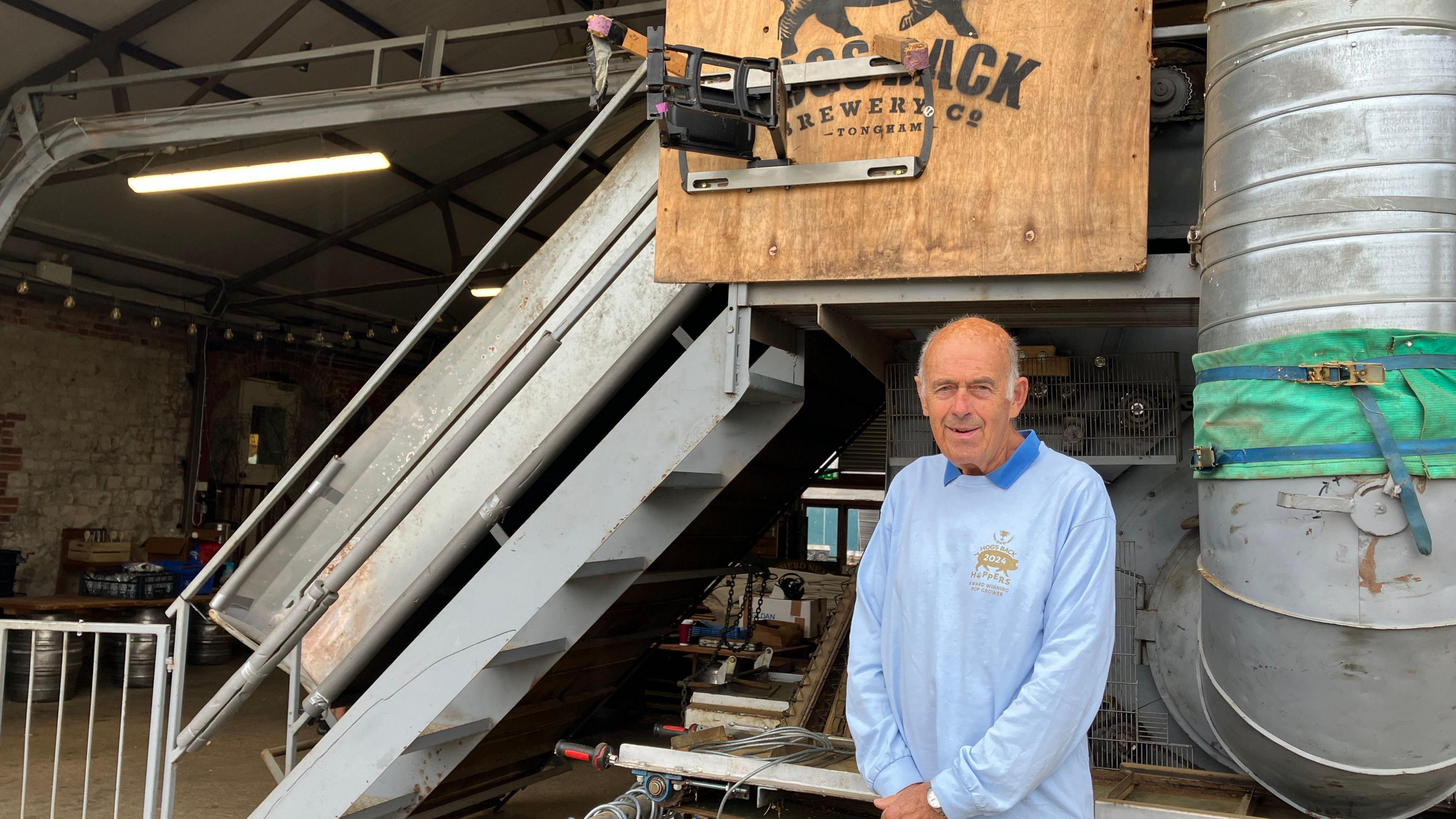 A picture of volunteer Ian Dixon, stood in front of machinery at the Hogsback Brewery in Tongham, Surrey. Ian is wearing a pale blue top. 