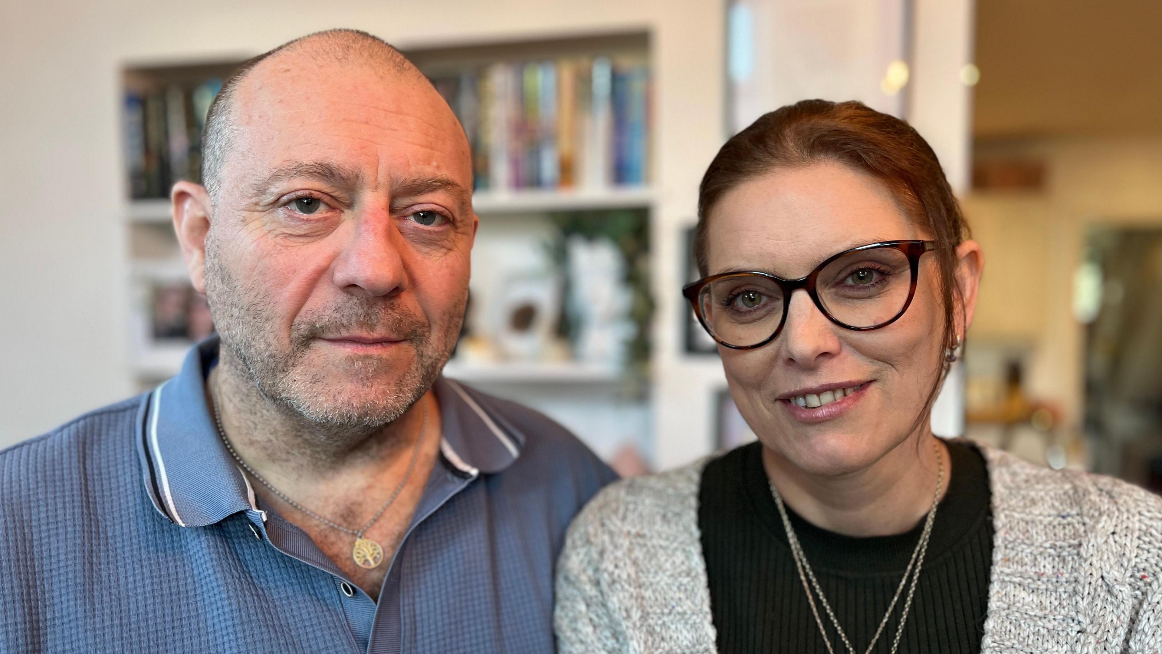 On the left Karl has short grey hair and a stubble beard and wears a blue polo shirt. On the right, Emma has dark hair tied back, black-rimmed glasses and wears a black jumper, light grey cardigan and two gold chain necklaces.