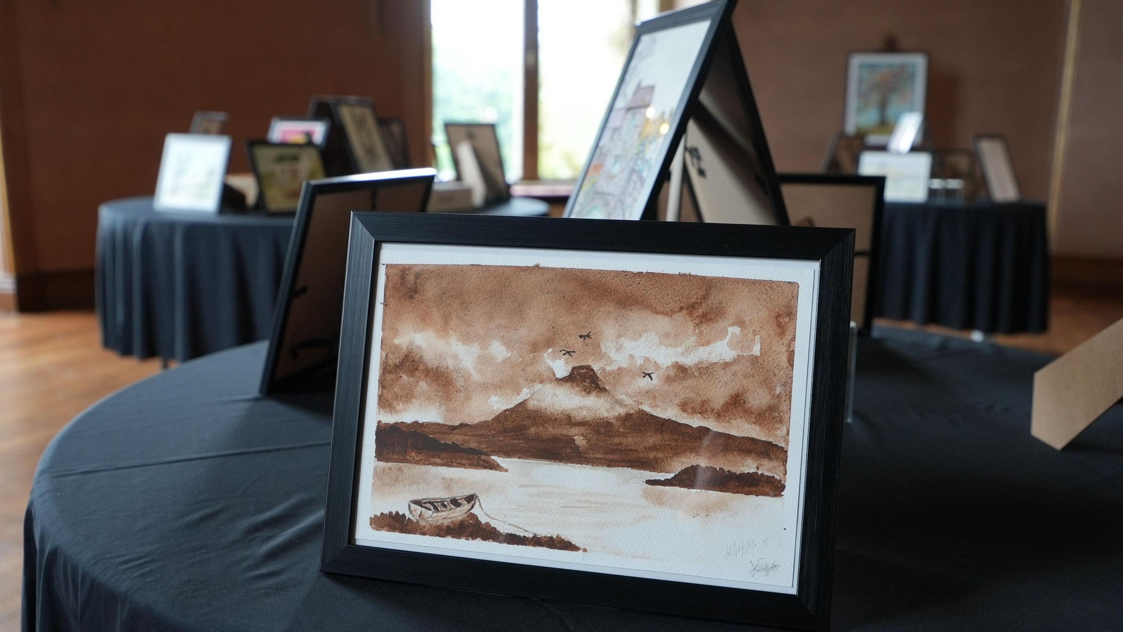 A watercolour painting of an island with a hill and a moody sky in colours of sepia and brown in a black frame. It is on a table with a black cloth with other paintings behind it and on other tables in the background. It is a large room with a window in the background.