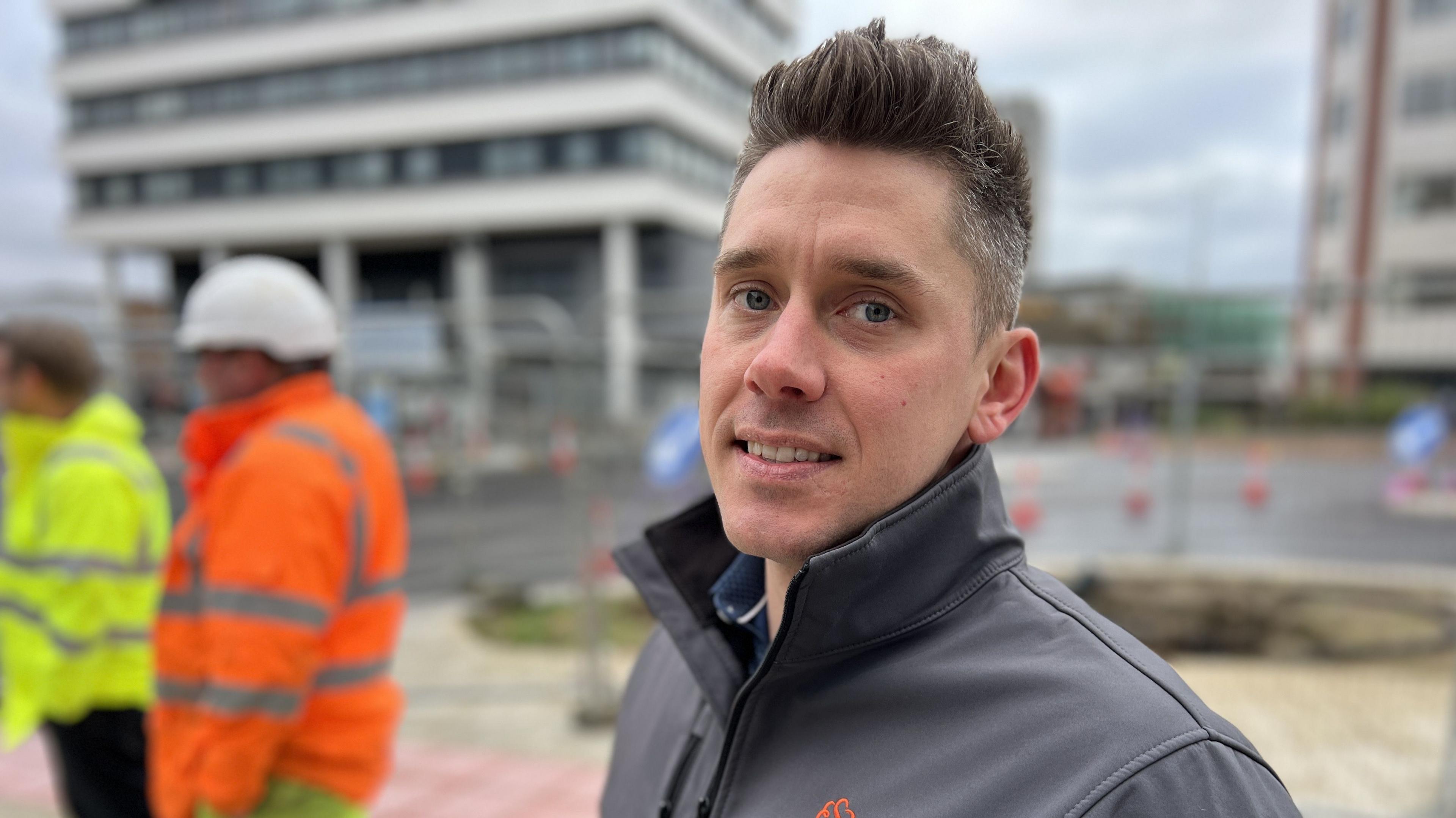 Bus Boulevard project manager, Liam Hill-Davis, with short cropped hair standing in front of workers with high-visibility jackets and hard hats in a construction site