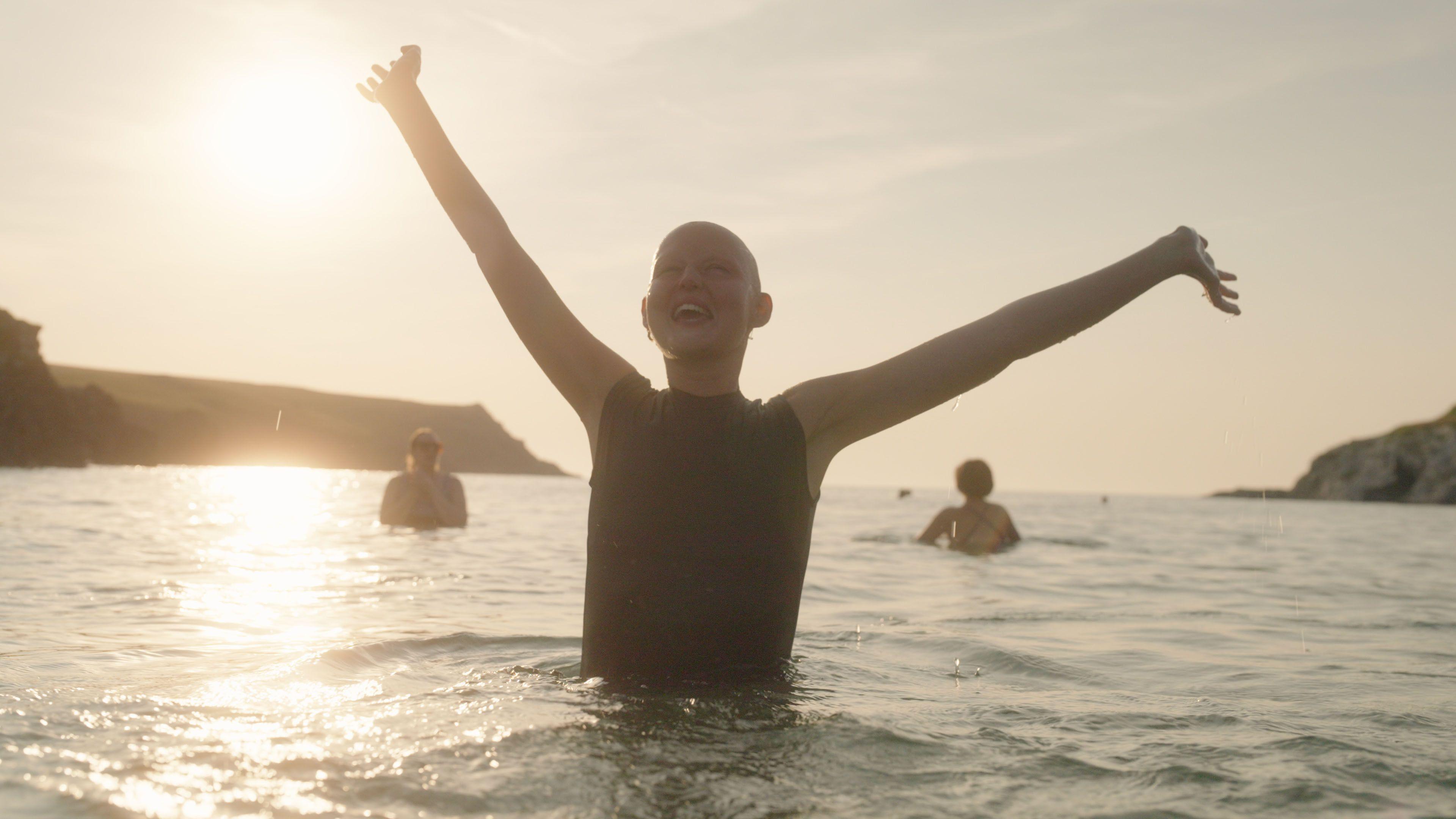 Kris is standing in the sea. She has her arms outstretched and is smiling. Two people can be seen in the background behind her and the sun is shining.